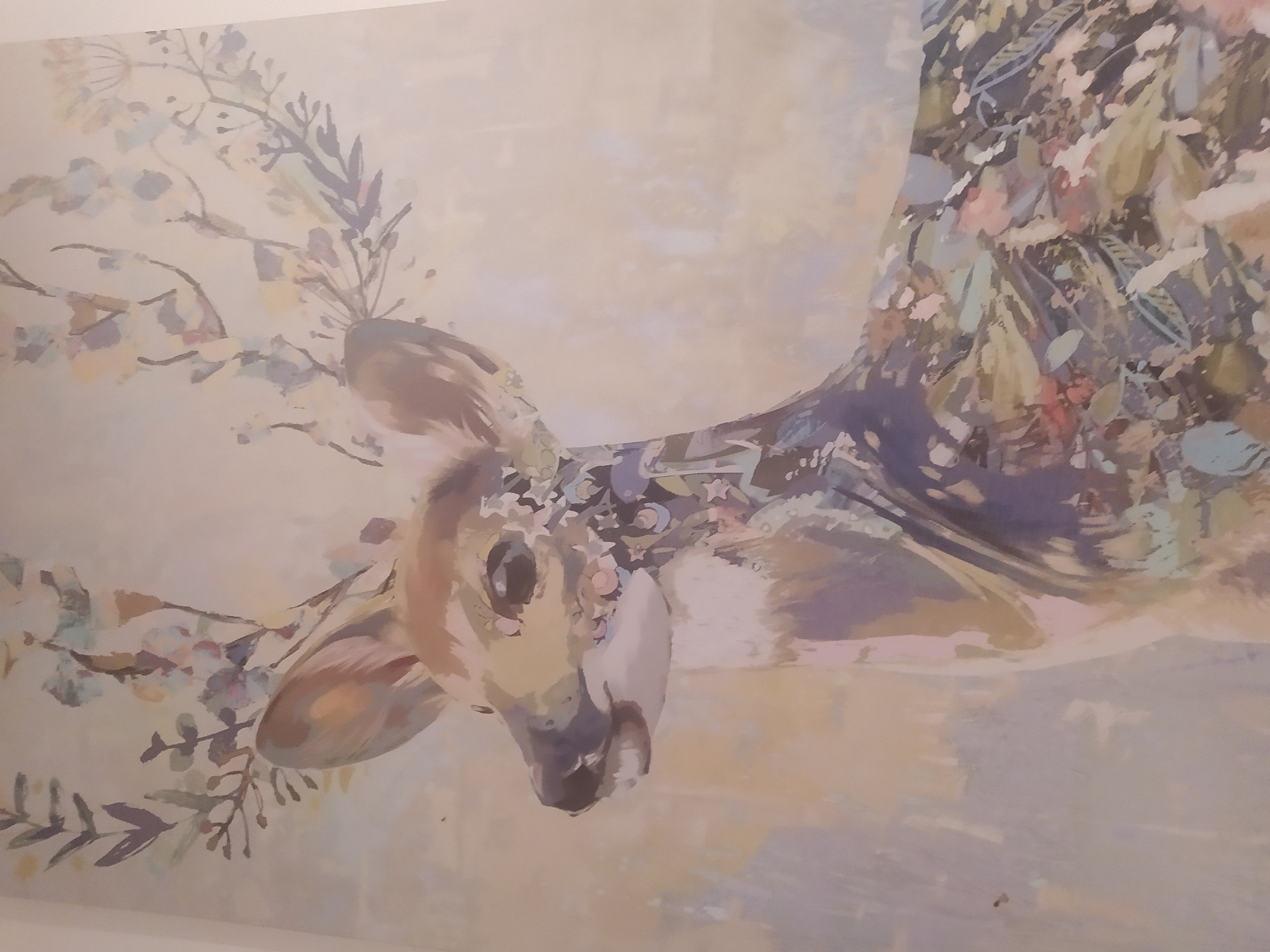 This is a close-up, eye-level, medium shot of a painting of a deer with floral antlers. The deer is facing left, with its head slightly lowered and its mouth open. Its body is covered in colorful floral patterns. The background is a soft, pale wash of color, with some blurry shapes that may represent trees or foliage. The lighting is even and diffused, suggesting the image was taken in a well-lit indoor space. The painting is rendered in a loose, impressionistic style with visible brushstrokes.