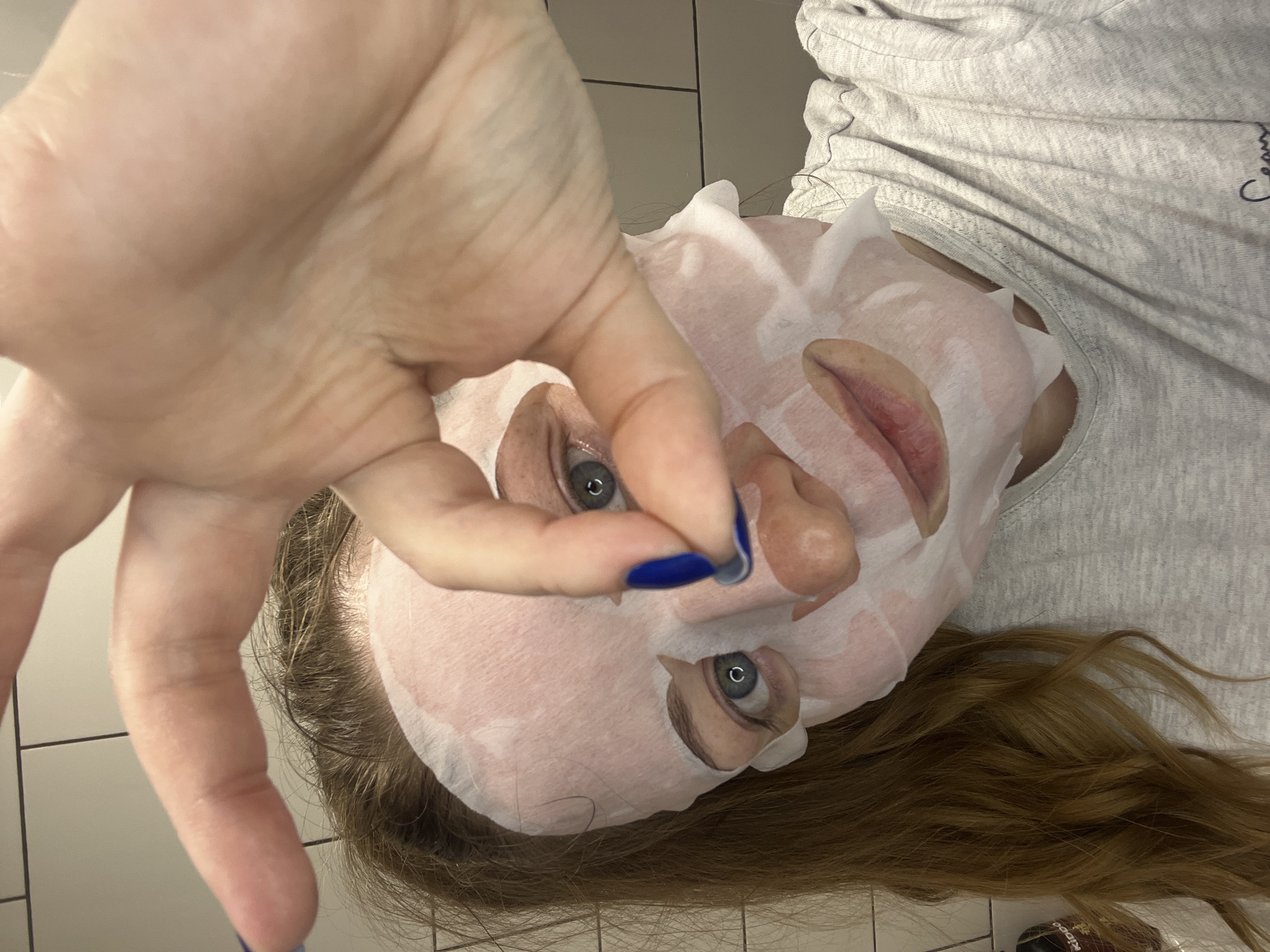 A woman is captured in a close-up, top-down perspective, engaging in a self-care routine with a sheet mask on her face. Her left hand, adorned with vibrant blue nail polish, is extended towards the camera in the foreground, creating a dynamic, selfie-like composition. Her face is mostly covered by a white, translucent sheet mask, which has cutouts revealing her light blue or grey eyes, nose, and lips. Her expression appears calm and relaxed, indicating a moment of personal indulgence.
She has long, flowing brown hair with auburn undertones, which spreads out around her head against a light-colored, possibly tiled, surface. She is wearing a light grey, possibly heathered, t-shirt. Partial text, "Cream," is visible on the fabric of her shirt near her shoulder. In the bottom right corner of the frame, partially obscured by her hair, a dark object with the word "KIDDO" printed on it can be seen, possibly a cosmetic product or container.
The setting appears to be indoors, likely a bathroom or personal space, suggested by the tiled background and the nature of the activity. The lighting is even, suggesting indoor artificial light. The image conveys a serene and intimate moment of self-care. This scene takes place in Раздолье, Russia.