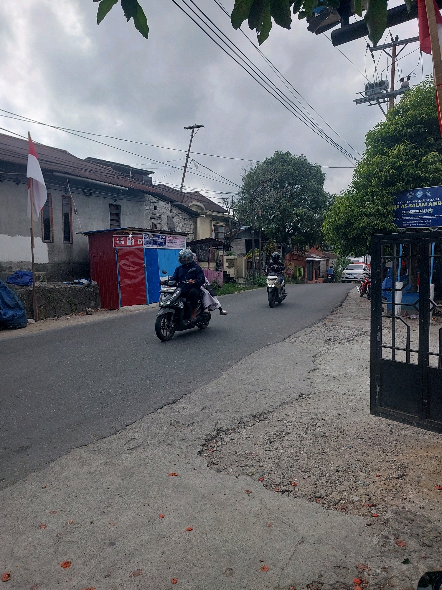 This is an eye-level, medium shot of a street in Ambon, Indonesia, on a cloudy day. Two motorcycles are seen on the road. The first motorcycle, in the foreground, is a dark scooter being ridden by a person wearing a dark jacket and a helmet. The second motorcycle, further down the road, is also being ridden by a person wearing a helmet, and it appears to be a lighter colored scooter. On the left side of the street, there are several low-rise buildings. One building has a red and blue facade, and a small sign that says "POM BENSIN MINI". Next to it, a white Indonesian flag is attached to a pole. On the right side of the street, there is a large gate and a sign that reads "YAYASAN AS-SALAM MALUKU MA AS-SALAM AMBON TERAKREDITASI". Trees are visible behind the sign. The road is paved, and the sidewalk on the right appears to be made of concrete and gravel. The sky is overcast, suggesting a possibility of rain.