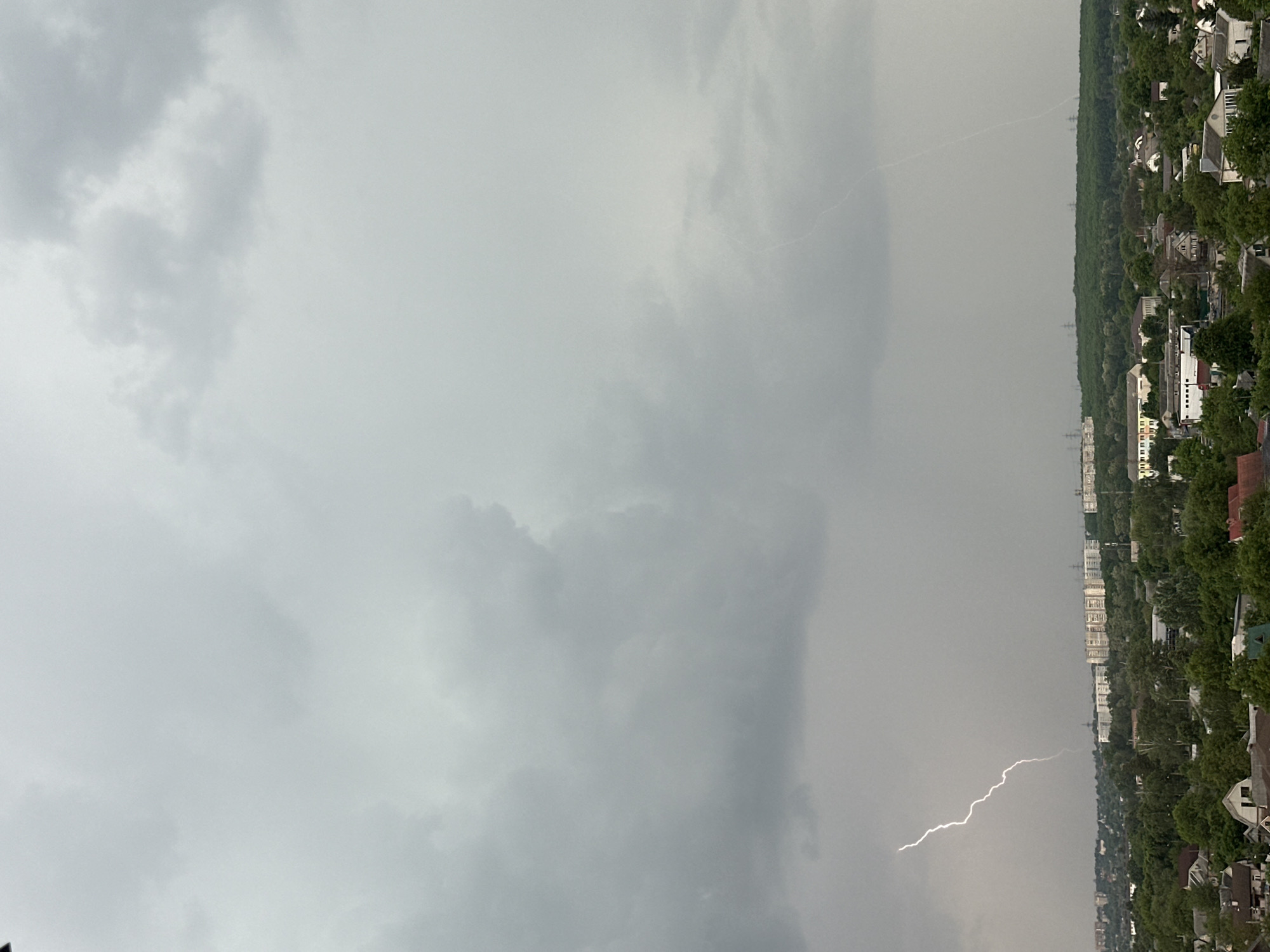 A sideways aerial view of a thunderstorm in an urban area. The sky is a dull gray, with thick clouds covering most of the frame. Lightning streaks downwards toward the lower-right corner, juxtaposed against a city-scape with buildings and tree-lined streets. The location is Unknown, Unknown.