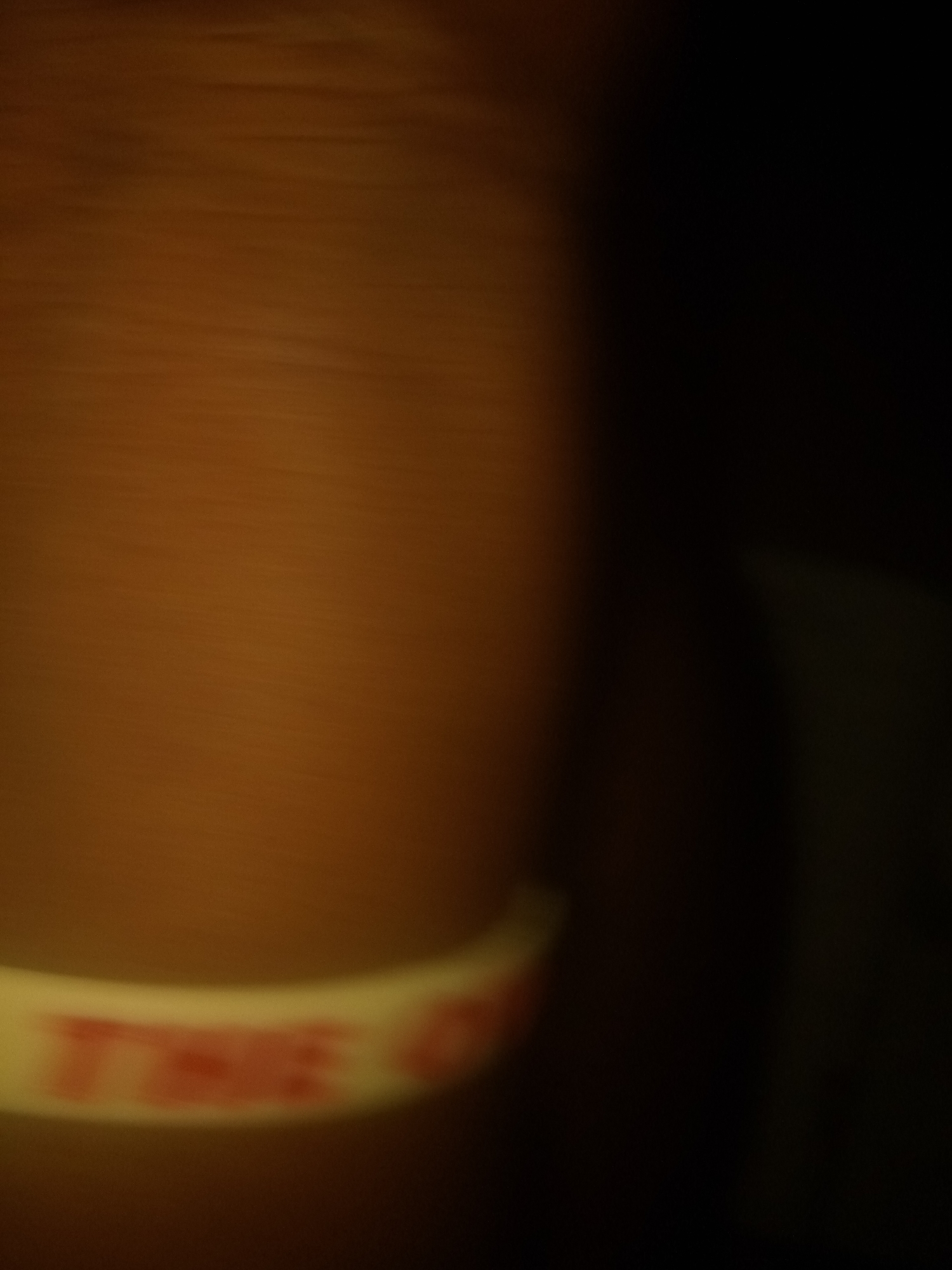 The image shows a blurred close-up of a white wristband with red lettering. The lettering is partially obscured by the blur, but the letters "TME" are visible, suggesting it might be part of a word like "TIME" or a brand name. The wristband is curved, indicating it is worn on a wrist, but no person is clearly visible. The background is dark and indistinct, with a warm, brown hue dominating the illuminated portion. The overall impression is of a dimly lit environment. There is no discernible information within the image to place it in the city of Jauro Yinu, Nigeria.