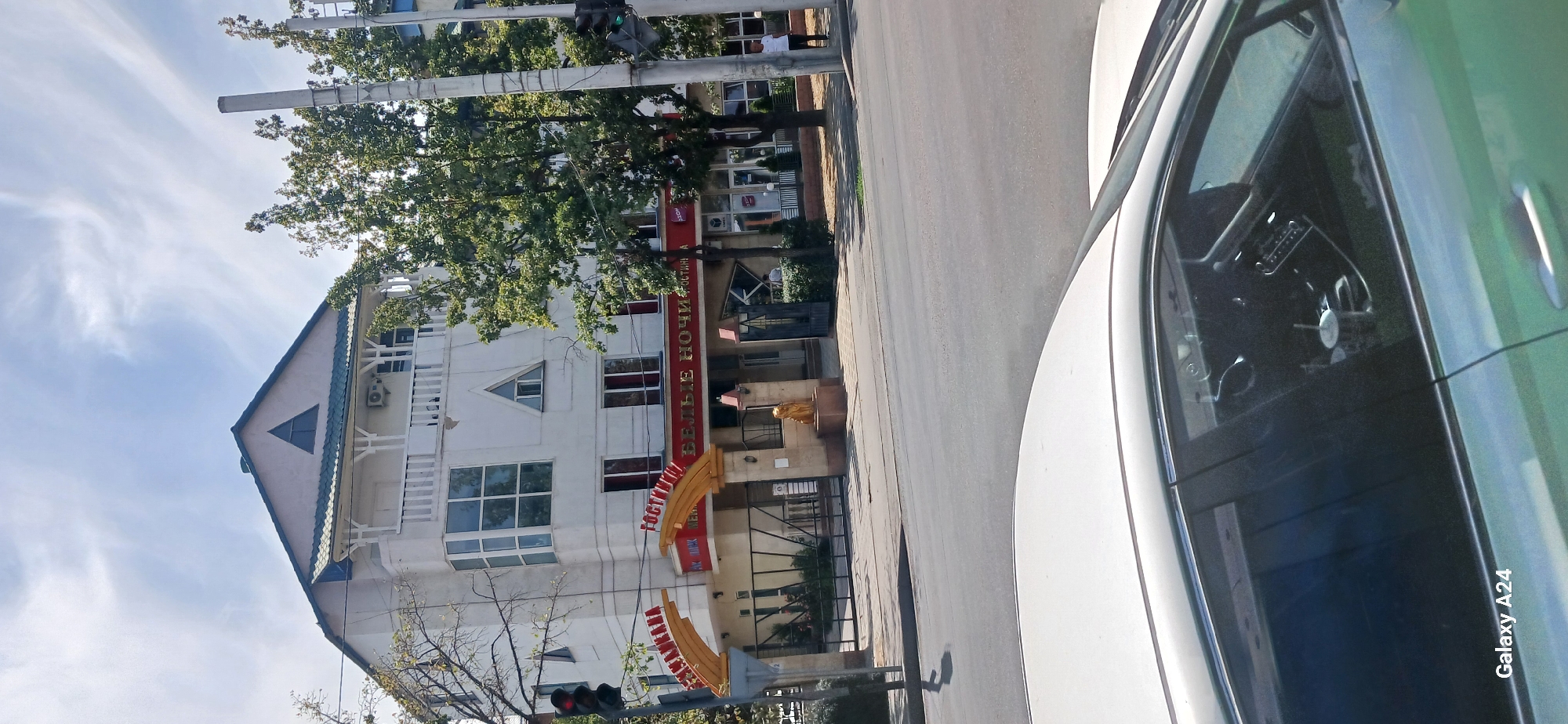This image shows a street view of a hotel building with a red sign that reads "ГОСТИНИЦА БЕЛЫЕ НОЧИ" (Hotel White Nights). The hotel is a white, multi-story building with a green roof and a balcony on the second floor. In front of the hotel, there is an entrance with a golden archway. A golden statue of a lion is visible near the entrance. The street is paved, and there are trees lining the sidewalk. A traffic light is visible on the left side of the image. In the foreground, the hood and windshield of a white car are visible. The sky is blue with some white clouds. The image appears to be taken in Bishkek, Kyrgyzstan, as indicated by the "Galaxy A24" watermark, which is a model of a Samsung phone, a popular brand in Kyrgyzstan.