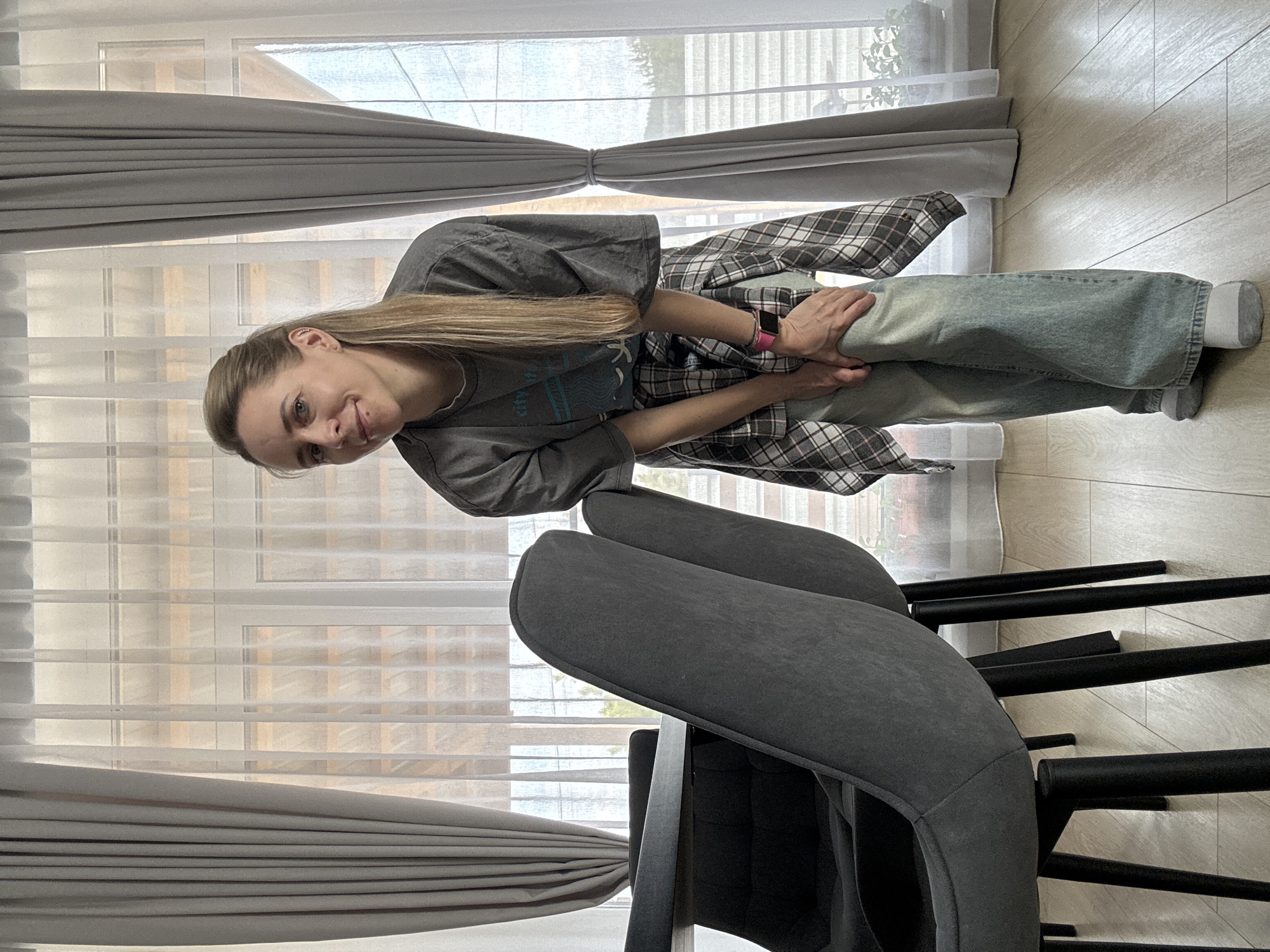 A smiling young woman with long blonde hair, pulled back, stands indoors, looking towards the camera. She is wearing a grey short-sleeved t-shirt with a light blue graphic and barely legible text, light blue jeans, and white socks. A plaid long-sleeved shirt is tied around her waist. A pink smartwatch is visible on her left wrist. Her hands are clasped in front of her, holding her jeans.
The setting appears to be a modern interior space, possibly a living or dining area. Behind the woman, a large window spans much of the wall, letting in natural daylight. Sheer white blinds or curtains cover part of the window, with grey opaque curtains drawn to the sides, tied back with fabric straps. Through the window, hints of green foliage and an outdoor structure are visible, suggesting a clear daytime.
In the foreground and to the right, two dark grey upholstered chairs with black metal frames are partially visible, suggesting a dining or seating arrangement. The floor is covered with light-colored, wood-patterned planks. The overall mood conveyed by the woman's smile is pleasant and relaxed.