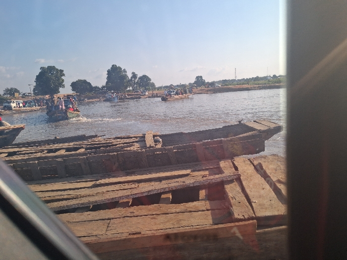 This image captures a busy waterfront scene in Nemnei, Nigeria, under a clear, light blue sky. In the immediate foreground, a weathered wooden pier or dock, constructed from rough planks, extends across the lower portion of the frame. The bow section of a dark, old wooden boat is partially visible alongside this structure. The view is partially obstructed by a dark, diagonal element in the lower left and a vertical dark object along the far right edge, suggesting the photograph was taken from within an enclosure.
The central area of the image is dominated by a body of water, likely a river, upon which multiple wooden boats are active. A prominent green motorboat, visibly overloaded with approximately 15-20 individuals, is moving from left to right, leaving a distinct white wake in its path. Other wooden boats are distributed across the water, some appearing stationary near the distant bank.
The far bank features a dirt shoreline, several large green trees, and a collection of parked vehicles, including white vans, indicating a land-based connection point. A faint communication tower is visible in the far background against the clear sky. The scene depicts a functional water transport hub.