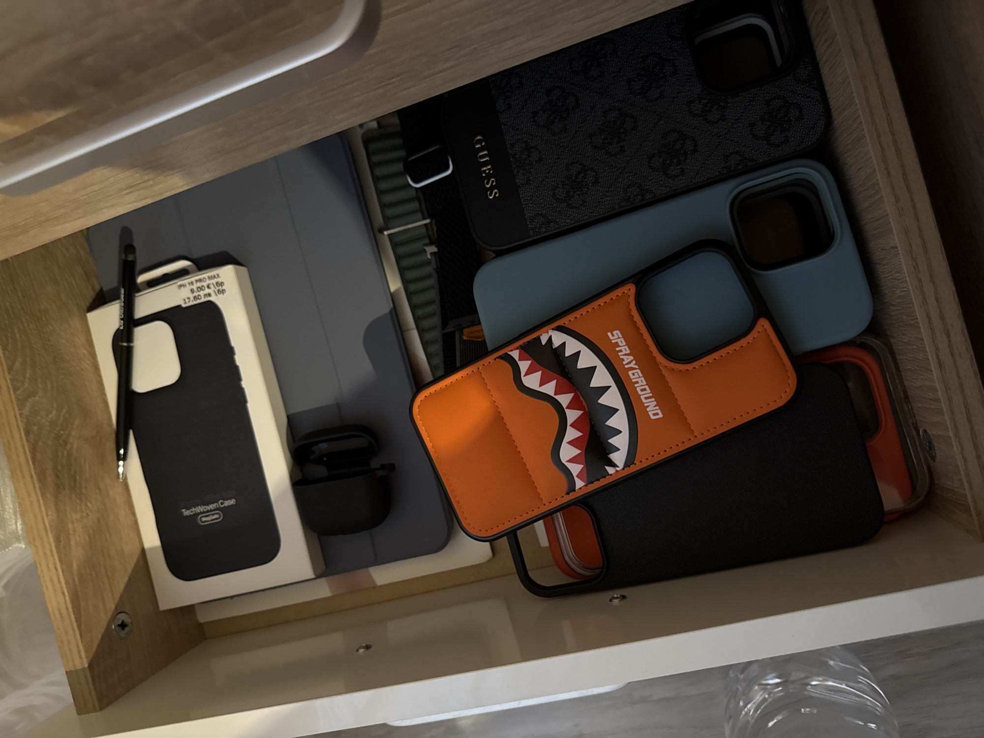 Here's a detailed description of the image:
The image shows an open drawer, possibly in a home setting. Inside the drawer are several items:
* **Phone Cases:** There are multiple phone cases visible. These include a black Guess case, a blue case, an orange "Sprayground" case, and a black "TechWoven Case" in its original packaging, which is labeled for an "IPH 10 PRO MAX" and priced at "9.00 €1бр" and "17.60 лв бр".
* **Tablet:** A blue tablet is partially visible.
* **Apple Pencil:** A black Apple Pencil is lying on top of the box of the TechWoven case.
* **Wireless Earbuds:** A set of black wireless earbuds are in the back of the box of the TechWoven case.
The scene appears to be within a piece of furniture, likely a dresser or nightstand, made of wood. The inside of the drawer is white.
It is impossible to determine the location in the city of Lovech, Bulgaria, or make further inferences about actions or interactions without additional information.