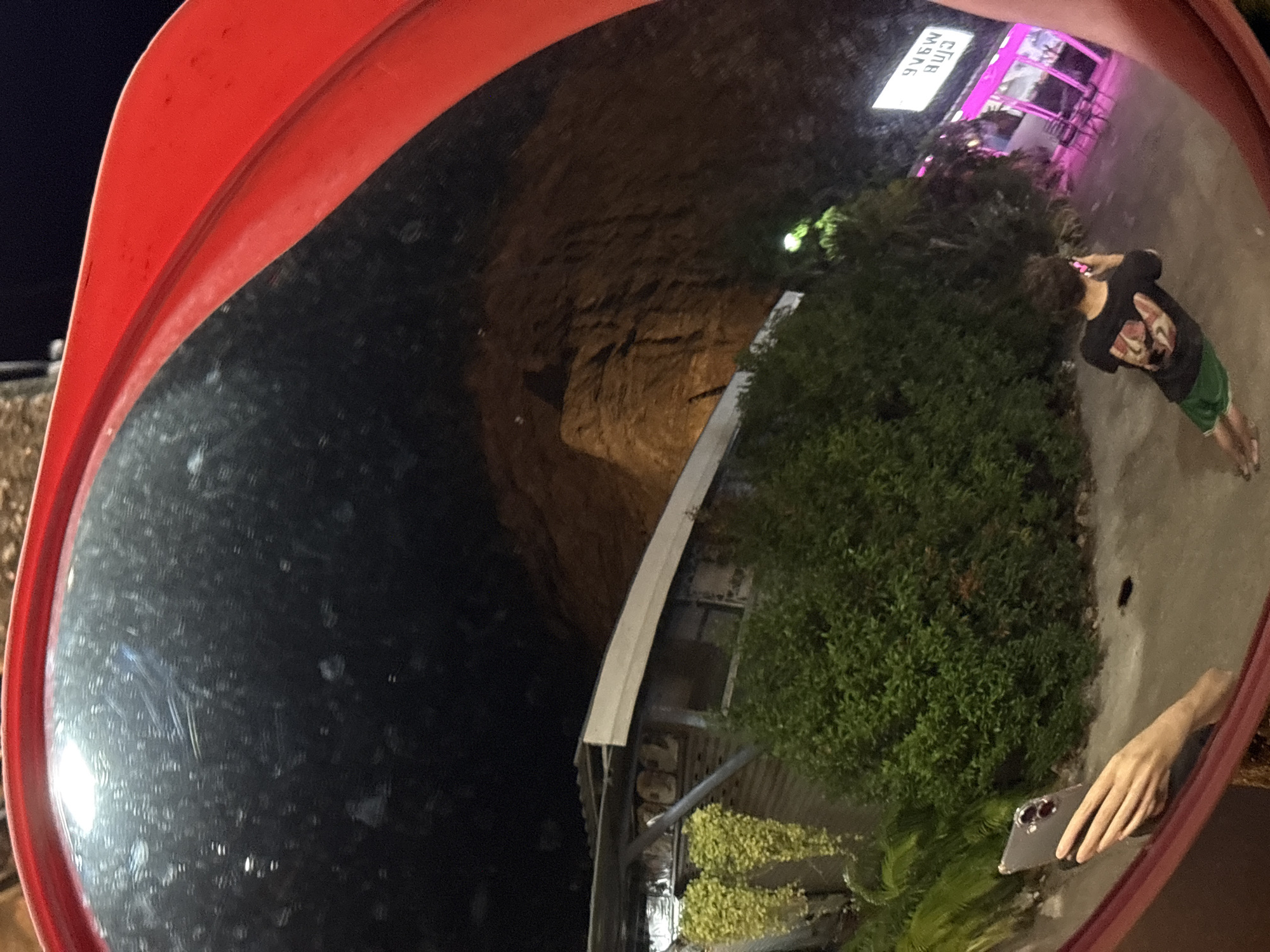 The image shows a convex mirror, with a red frame, reflecting a street scene in Baan Bang Rak, Thailand, at night. The reflection captures a man in a dark T-shirt and green shorts standing on the pavement near a building with a sign that says "CLUB M9A6." Adjacent to the man is a large, green bush. In the lower right corner of the mirror's reflection, a hand holding a smartphone is visible. The left side of the mirror's reflection depicts a dark area, possibly a rock formation or hillside. The overall scene is dimly lit, with some brighter lighting near the building with the "CLUB M9A6" sign.
