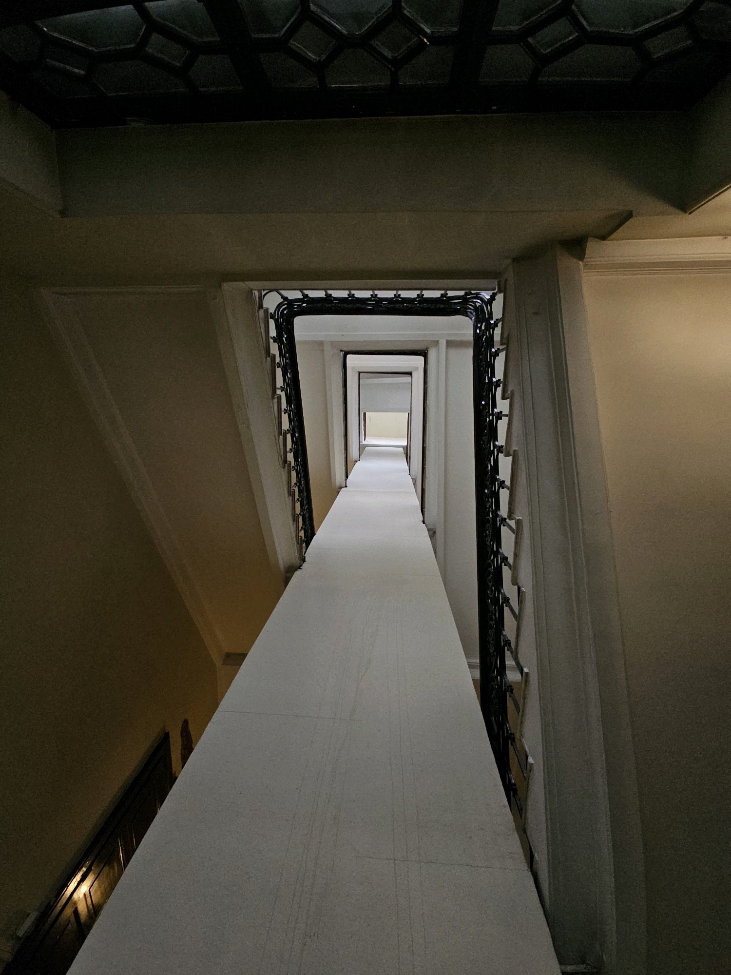The image shows a perspective view looking down a long, narrow, dimly lit corridor or staircase. The dominant features are a white, seemingly stone or concrete banister running diagonally from the foreground to the background, and a dark, ornate metal railing on the right side. The walls on either side of the corridor are painted in a light, neutral color and show some architectural molding. The ceiling at the top of the image has a dark, geometric pattern. The corridor recedes into the distance, with multiple doorways or archways visible, creating a sense of depth and repetition. The lighting is uneven, with some areas brightly lit by an unseen source, and others in shadow, contributing to a somewhat mysterious or atmospheric quality. The scene appears to be an interior architectural space, possibly a building with historical elements. There are no people or any obvious signs of recent activity. The image does not contain any discernible elements that would place it specifically in Moscow, Russia.