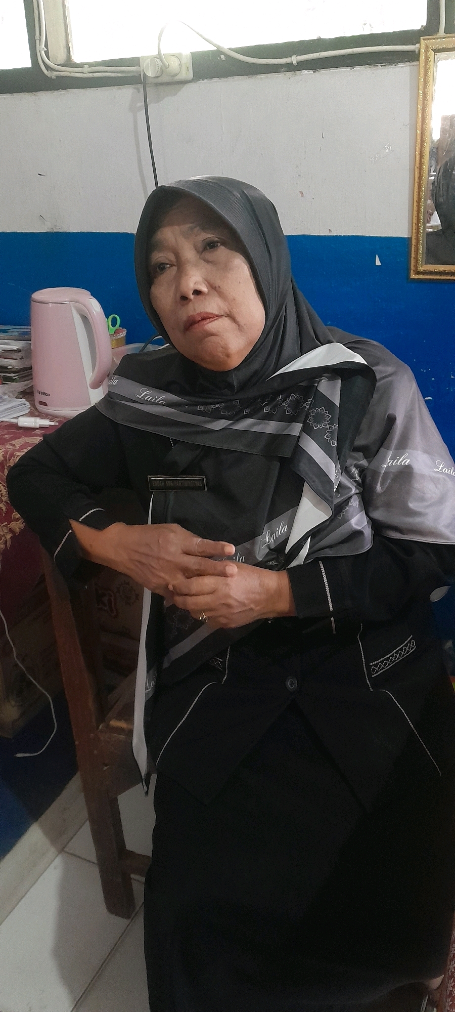 The image shows an elderly Indonesian woman wearing a black hijab and a black uniform with a grey and white patterned scarf. She is seated in what appears to be an indoor setting, possibly an office or a home, with a blue and white wall in the background. A pink electric kettle is visible on a table to her left, along with some papers and a pair of scissors. A framed mirror hangs on the wall to her right. The woman's hands are clasped in her lap, and she is looking slightly away from the camera. The scene suggests a formal or professional environment, common in offices or government buildings in Indonesia, such as those found in the city of Tahunan, Indonesia, where the administrative offices for the Jepara Regency are located.