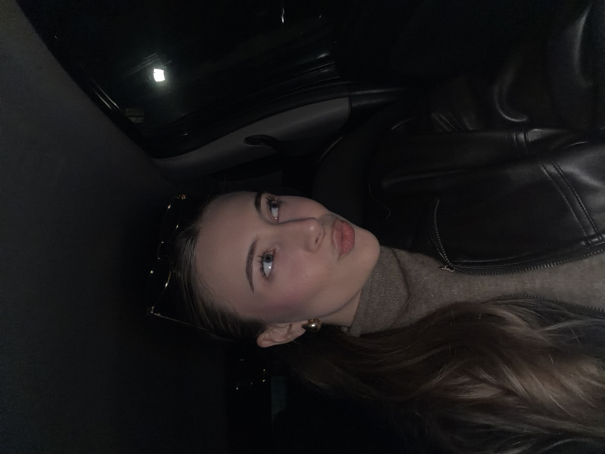 A young adult female is seated in the front of a vehicle, possibly the driver's or front passenger's seat, at night. She is positioned towards the left side of the frame, looking to her right with a puckered lip expression. Her long, light brown hair is styled in a ponytail to her left side. She wears a light brown turtleneck sweater and a dark, likely black, leather jacket. Accessories include gold-toned hoop earrings, a small stud earring on her visible ear, a thin chain necklace, and dark-framed sunglasses perched on top of her head. The vehicle's interior is dark, with the passenger-side A-pillar and a portion of the door frame visible on the right. A distinct, bright white, rectangular light source is reflected in the car's side window, suggesting an external streetlamp or building illumination. The exact location within the city of Plovdiv, Bulgaria, cannot be determined from the visual information present in the image.