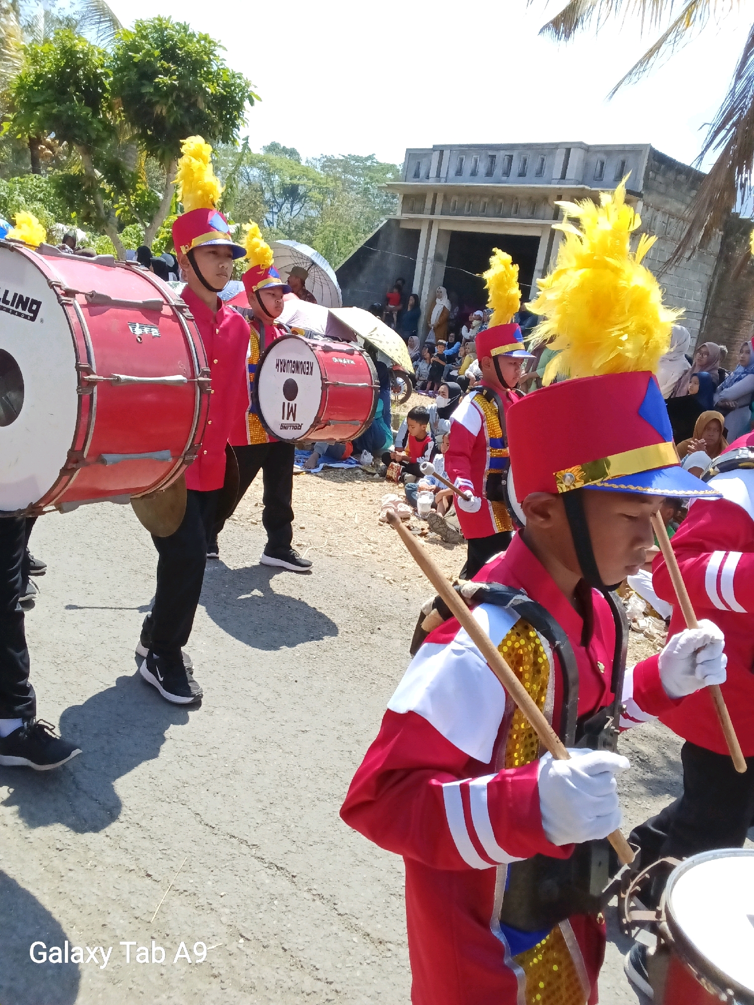 A group of young boys in red and blue marching band uniforms with yellow feathered hats are parading in the street. The boy in the foreground is wearing a red jacket with white accents and a blue trim on his hat, holding drumsticks. He is positioned to the right of a large red bass drum with a white "LLING" logo on its side. To the left of the bass drum, another boy in a similar uniform is visible. Behind them, other band members and spectators are present. The background features a building with a gray facade, palm trees, and more onlookers. The ground is paved with asphalt. The image is taken outdoors under bright sunlight. A watermark at the bottom left corner reads "Galaxy Tab A9".
