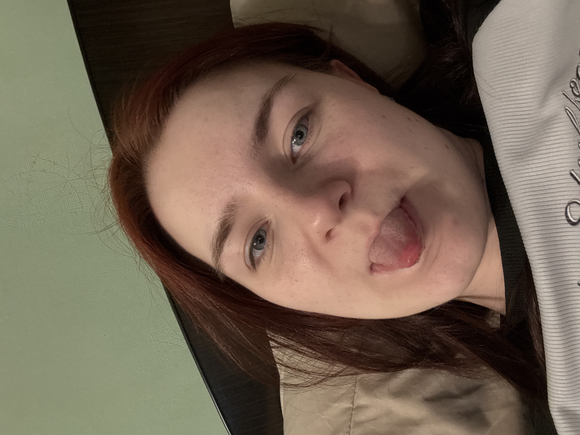A woman with reddish-brown hair is lying down and sticking out her tongue. She is wearing a light-colored ribbed shirt. Her face is close to the camera, and she is looking slightly off to the side. The background appears to be a bed with a light-colored pillow and a wall with a faint texture. The image was taken indoors in Sinyavino, Russia.