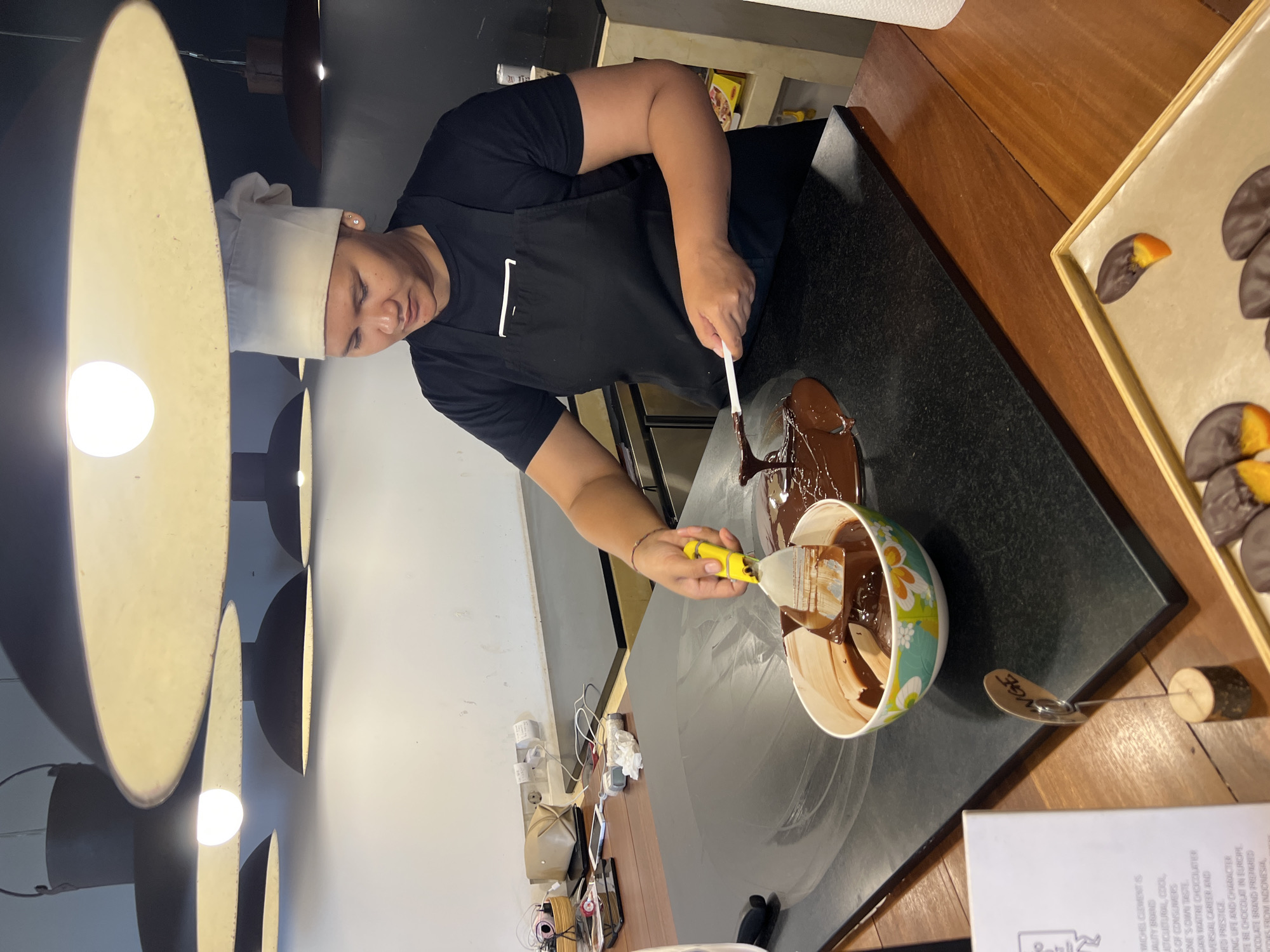 A chef is preparing chocolate treats in a brightly lit kitchen, likely in the city of Unknown, Unknown. The chef, wearing a black uniform and a white toque, is using a yellow and white spatula to stir melted chocolate in a floral-patterned bowl. Some of the chocolate has spilled onto the black countertop. Finished chocolate-covered treats, some with orange slices, are arranged on a tray to the right. Other kitchen equipment and supplies are visible in the background. A brochure about the ingredients of the chocolate is on the table, partially covered by the chocolate bowl.