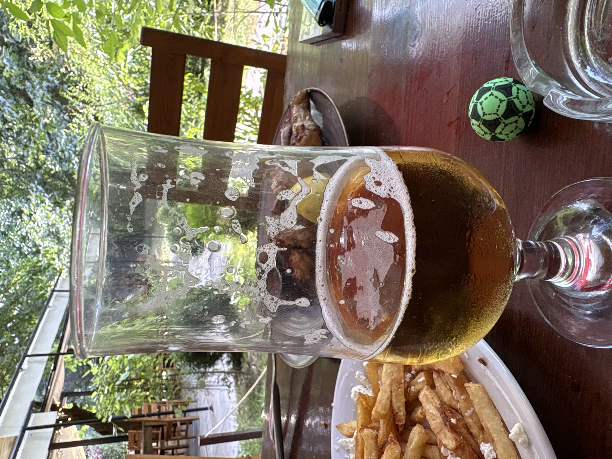 In Parvenets, Bulgaria, a half-empty glass of amber-colored beer sits on a dark wooden table. The glass has foamy residue clinging to its sides. Beside the glass is a plate of golden french fries, some topped with crumbled white cheese. In the background, a dark wooden chair is visible, and beyond that, lush green foliage and trees suggest an outdoor setting. A small, black and green soccer ball rests on the table to the right of the beer glass, next to an ashtray. The overall scene appears to be a casual dining or relaxation setting, possibly at a restaurant or outdoor cafe in the town of Parvenets.