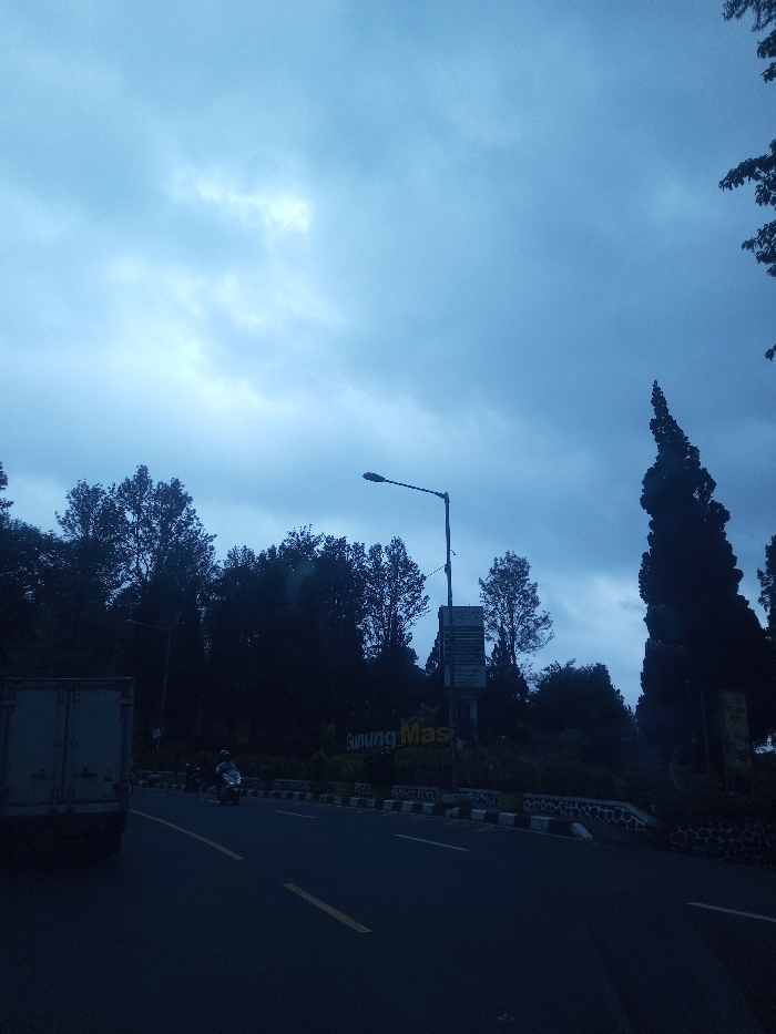 The image displays a road scene under an overcast, bluish-grey sky with a brighter patch visible in the upper left. The setting is identified by prominent signage reading "Gunung Mas," a name associated with locations in Indonesia. The asphalt road occupies the lower portion of the frame, featuring visible yellow double solid lines and white dashed lines. A white cargo truck is partially visible on the far left, moving away from the viewer. Further down the road, a single individual rides a white scooter or motorcycle, also moving away. The road is bordered by a black and white striped curb or median barrier. Dense, dark green trees line both sides of the road, appearing heavily shadowed. A tall, dark street lamp stands prominently towards the right-center, extending over the road. Below the street lamp, a rectangular sign post with multiple dark panels is visible. A distinct, tall, narrow coniferous tree is prominent on the far right. No specific interactions between individuals are discernible.