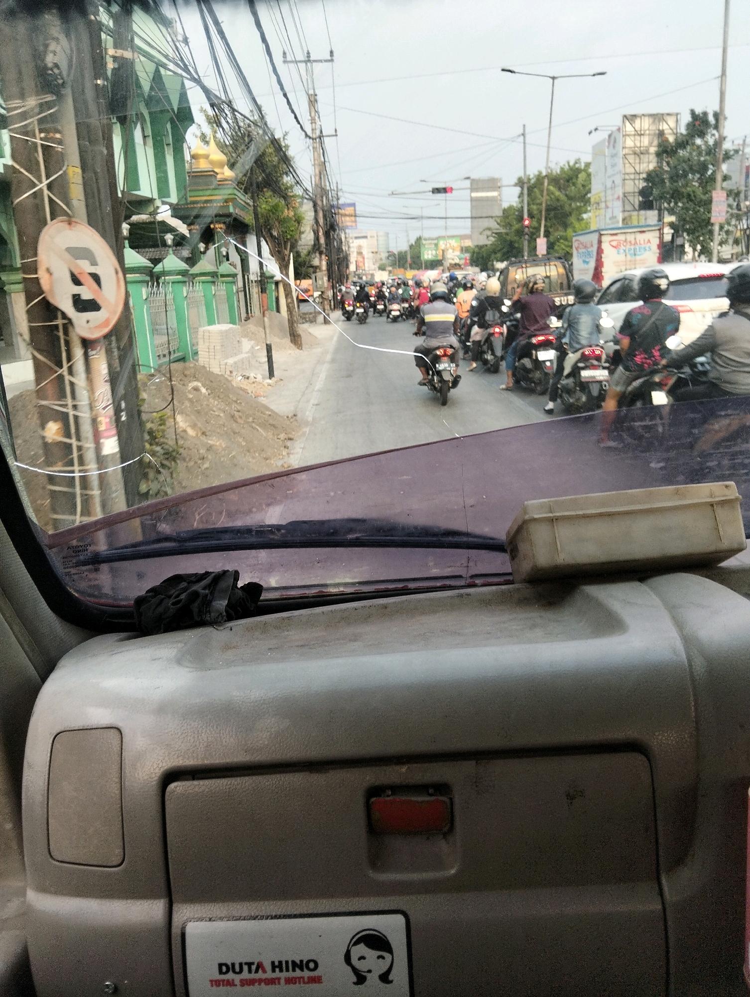 The image captures a street scene in Semarang, Indonesia, likely during daylight, depicting moderate traffic. A prominent green and gold mosque is visible on the left, with a "no stopping" sign in the foreground. The road is filled with motorcycles, with a few cars also present. The perspective suggests the photo was taken from inside a vehicle, looking forward through the windshield. The dashboard of the vehicle is partially visible, showing a beige plastic console and a black cloth on the windshield. In the background, tall buildings and advertisements suggest an urban environment. The sky is overcast, and electrical wires crisscross above the street.