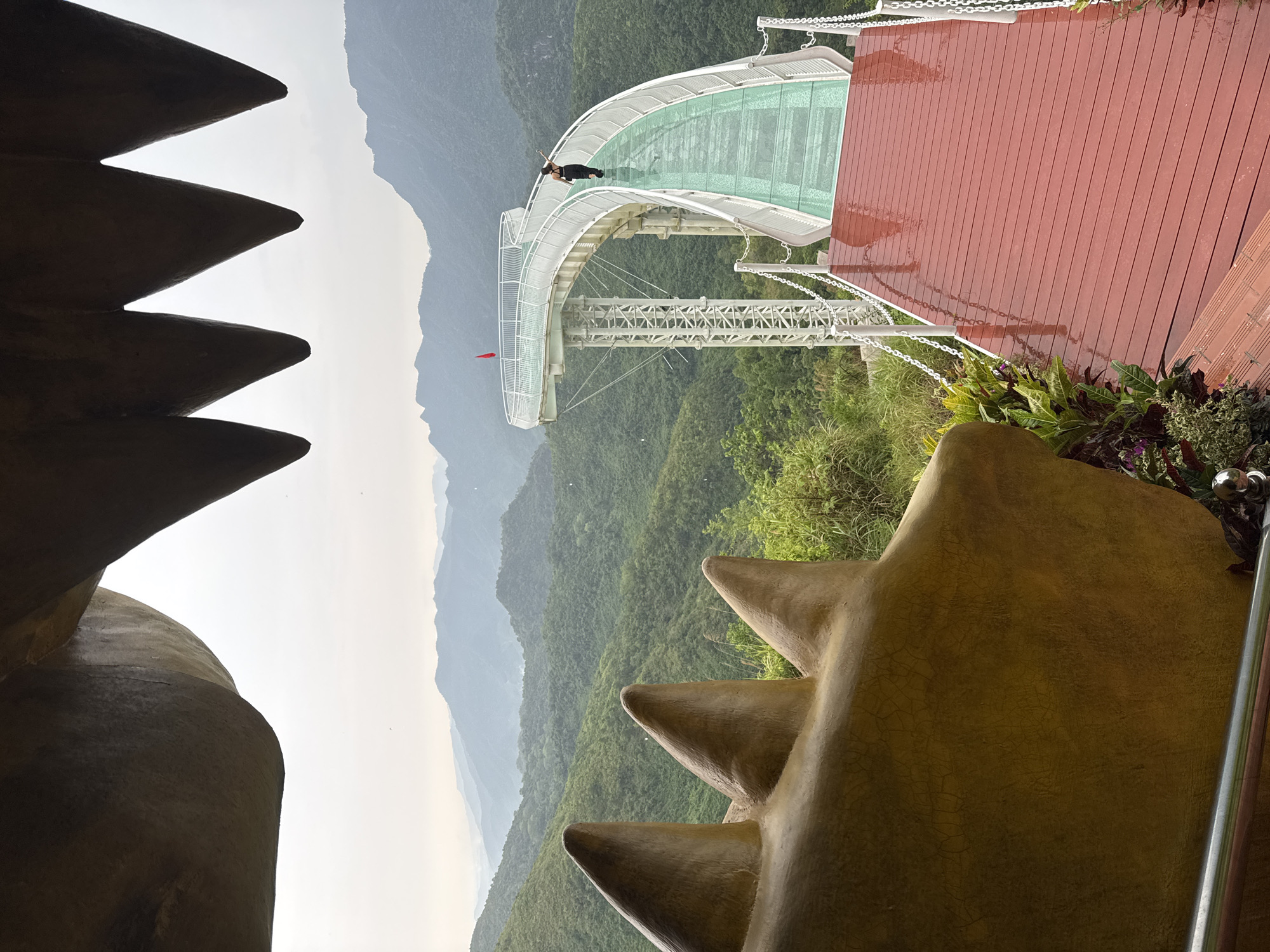 This image captures a dramatic architectural structure set within a vast mountainous landscape, likely a scenic tourist attraction in Đà Nẵng, Vietnam. The scene is dominated by a striking glass-bottomed bridge, which gracefully curves through the center of the frame. This bridge features white structural elements, railings, and a transparent, greenish-tinted floor.
One person, dressed in dark clothing, is visible on the glass bridge, lying down or leaning over to observe the view below, highlighting the bridge's unique transparent feature. To the right of the bridge, a wide reddish-brown wooden deck provides an observation platform, showing signs of recent rain or dew. Metallic chains serve as a barrier along the edge of this deck and connect to the bridge structure.
In the immediate foreground, large, stylized sculptural elements frame the view. On the left, a dark brown, serrated structure resembles giant teeth or spikes. On the right, a similar golden-brown, textured structure with pointed elements adds to the unique aesthetic, possibly depicting parts of a dragon or mythical creature.
Beyond the bridge and platforms, layers of lush green, forested mountains stretch into the distance, fading into a hazy, bright sky. A small red flag is visible near the far end of the glass bridge. The time of day appears to be daytime, with soft, diffused light, indicating either an overcast sky or perhaps early morning/late afternoon. The dampness on the wooden deck suggests recent precipitation. The overall scene conveys a sense of awe and adventure, inviting viewers to experience the panoramic vistas from this high-altitude viewpoint. No visible text is present in the image.