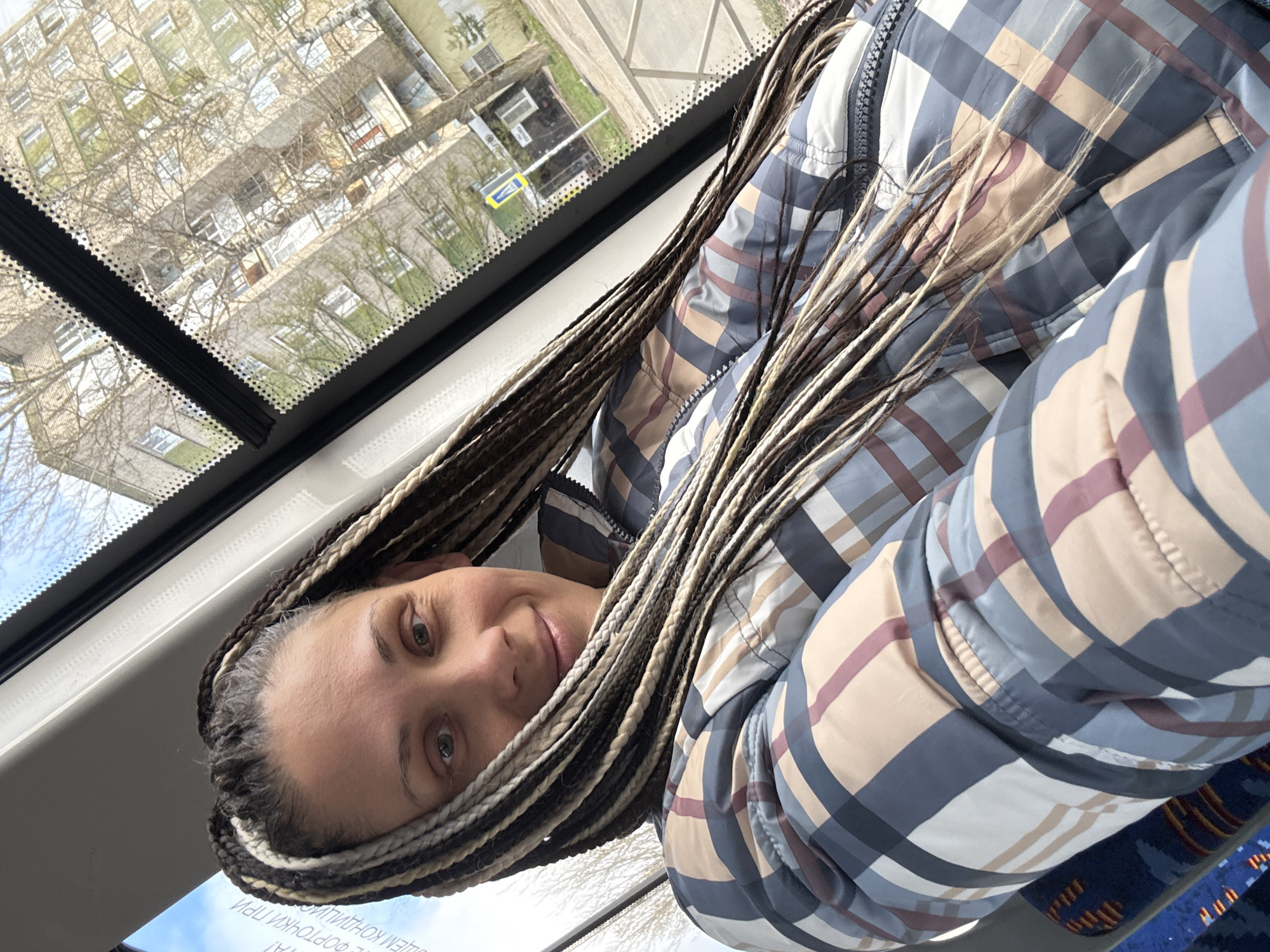 A woman with long brown and blonde braids is sitting on public transportation, likely a bus or trolley, visible through the window behind her. The buildings reflected are a mix of older apartment buildings, likely located in Rusanovo, Russia. The woman is wearing a blue, white, and tan plaid jacket, indicating it might be a cooler season, potentially spring or fall. The photo appears to be taken from a slightly low angle.