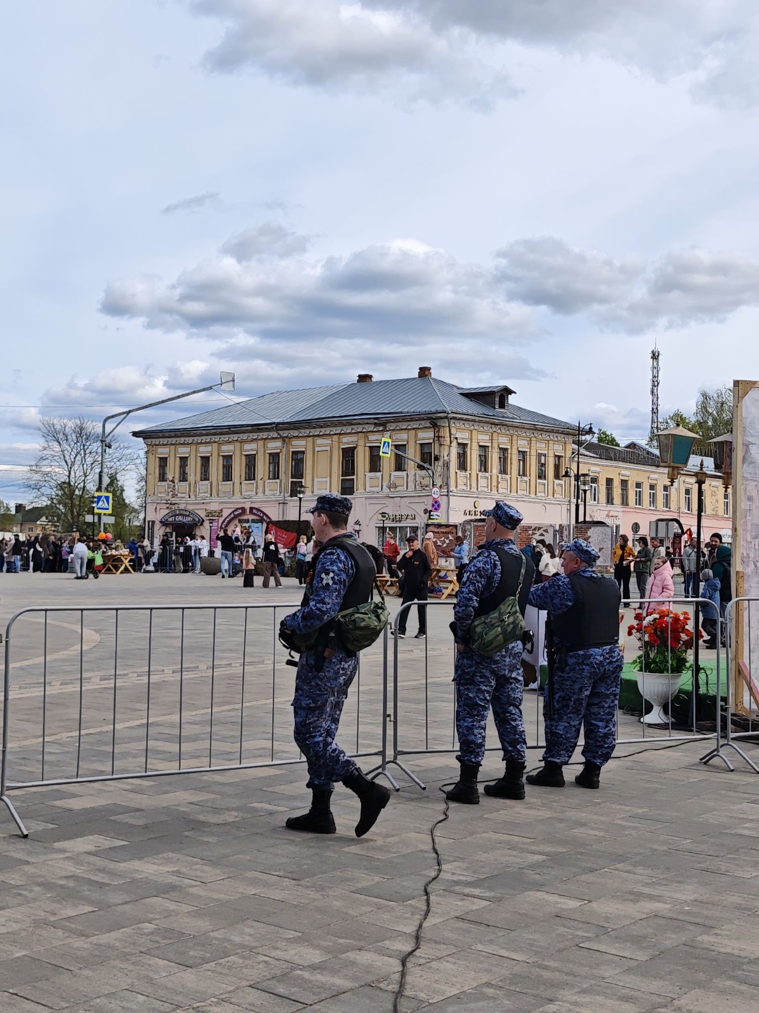 The image depicts a public space in what is possibly the city of Chelganovo, Russia. In the foreground, a metal barricade stretches across the frame, separating the viewer from the main activity area. Three uniformed individuals, possibly security personnel or police officers, stand behind the barricade. They wear camouflage uniforms with black vests and caps. One officer is walking, while the other two are standing.
Behind the barricade, a crowd of people is gathered in what appears to be a public event or market. There are vendor stalls, decorative elements, and general public activity. The architectural backdrop features a two-story building with a light-colored facade and a grey roof. A streetlight stands near the building. In the background, a cell tower is visible. The sky is overcast with grey and white clouds. The ground is paved with grey interlocking blocks. A tree with bare branches is also present on the left side of the frame.