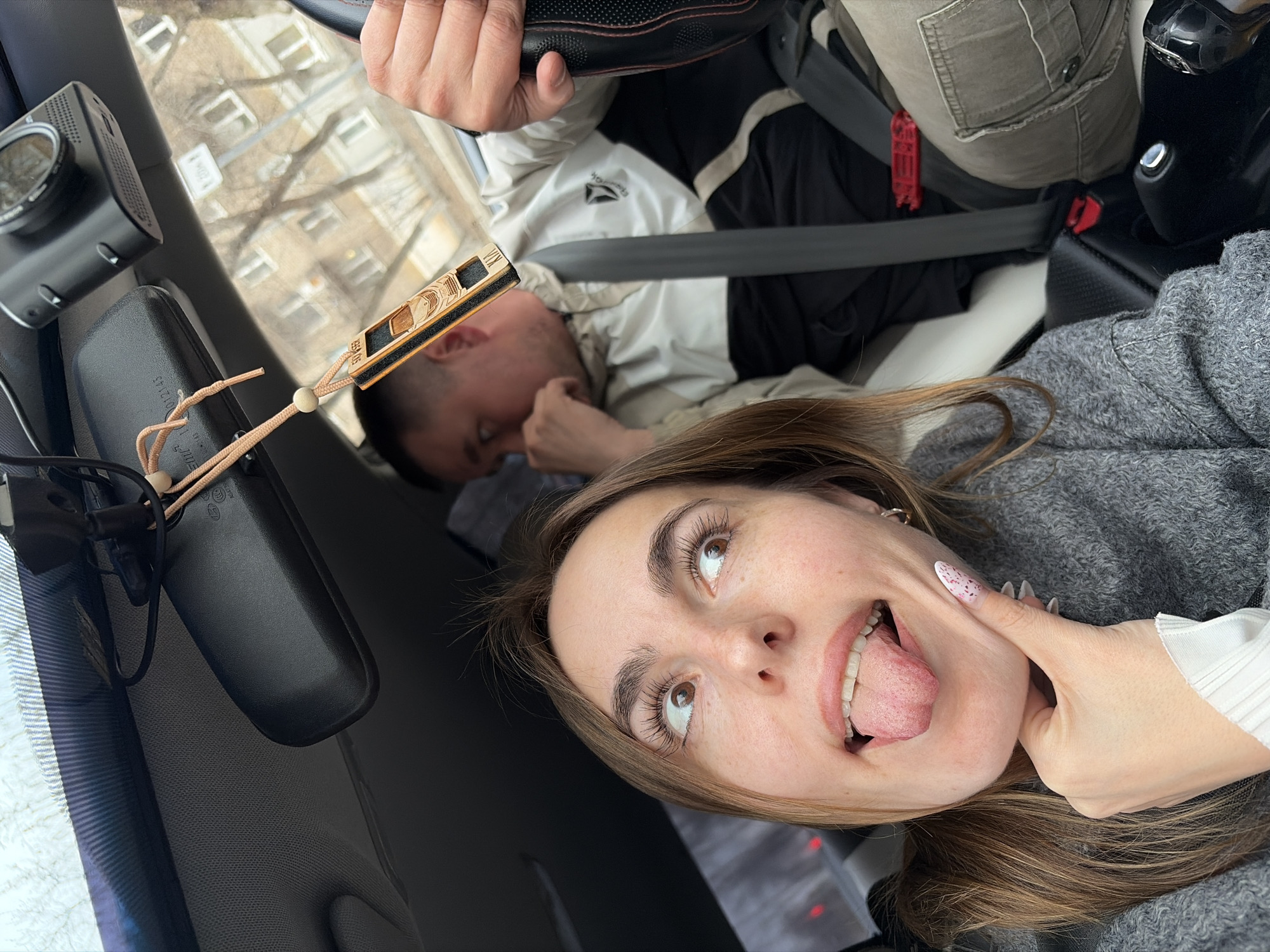 A selfie shot from within a vehicle shows two people and an exterior view of Izhevsk, Russia through the front windshield. The person taking the photo is a young woman with light brown hair, wearing a gray sweater and white top. She is sticking out her tongue and touching her cheek with her hand. To her left, in the passenger seat, is a man wearing a light-colored jacket and black pants, secured with a seatbelt. The driver is visible with his hands on the steering wheel. A rear-view mirror and dashcam are visible attached to the windshield. Outside the window, blurred buildings are visible, suggesting the location is in Izhevsk, Russia.