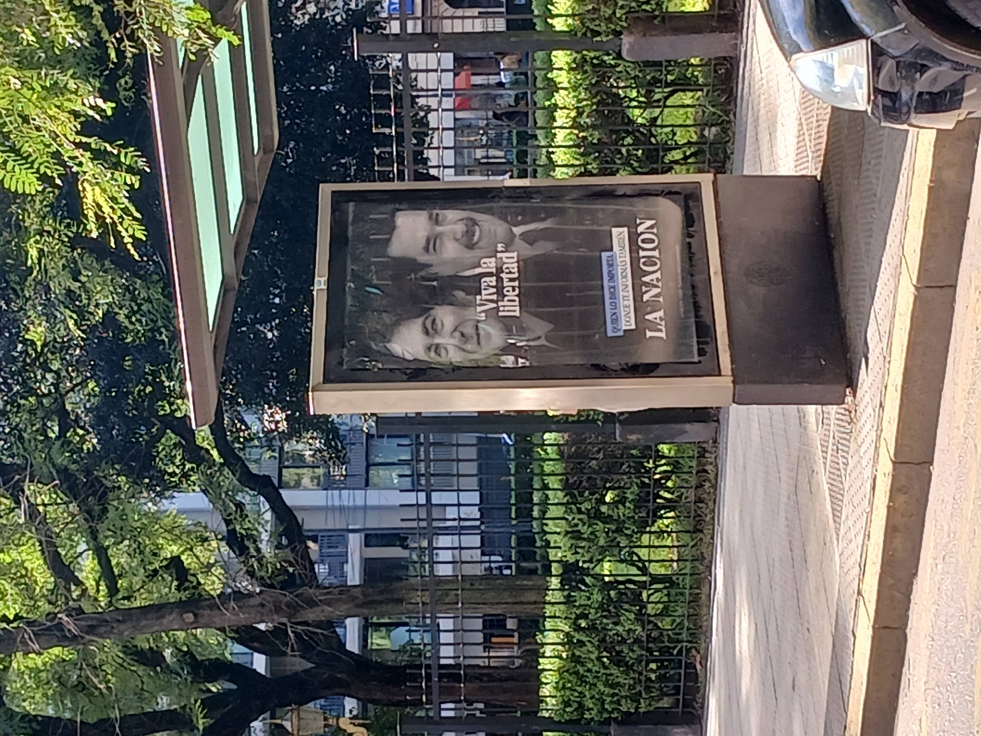 The image shows a sign for the Argentinian newspaper "La Nacion" located in Buenos Aires, Argentina. The sign is a metallic structure with a poster showing two smiling men along with the words "Viva la libertad." The sign is positioned on a sidewalk. Behind the sign, there is a green metal fence with dense vegetation visible. In the background, there is a multi-story building with many windows. Part of a vehicle is visible in the right edge of the frame. A tree is visible in the left portion of the image.