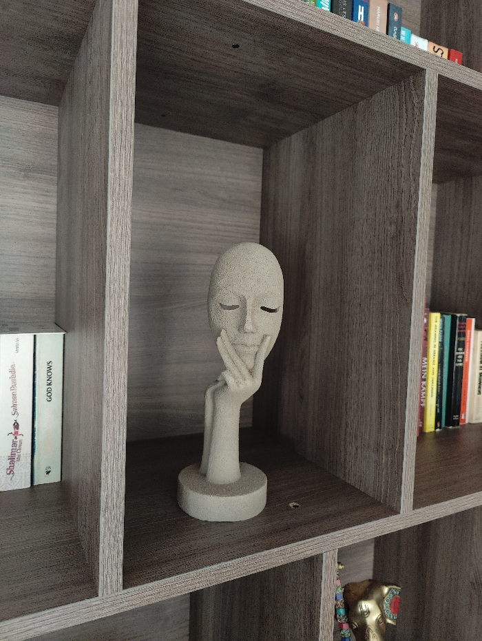 An indoor scene in Uithoorn, Netherlands, depicts a multi-compartment bookshelf constructed from light brown, wood-grain laminate. Centered within one compartment is a textured, light beige sculpture of an abstract human head with closed eyes and a serene expression, its right hand gently supporting the chin. The sculpture rests on a circular base. To the left of this compartment, several books with white spines are stacked horizontally, with titles like "GOD KNOWS" and "Salman Rushdie" partially legible. To the upper right, a vertical stack of books with various colorful spines (yellow, green, blue, red) is visible in another compartment. In the lower right compartment, a golden, ornate figurine, possibly an elephant head adorned with colorful beads, is partially visible. The environment appears organized, well-lit, and without human presence.