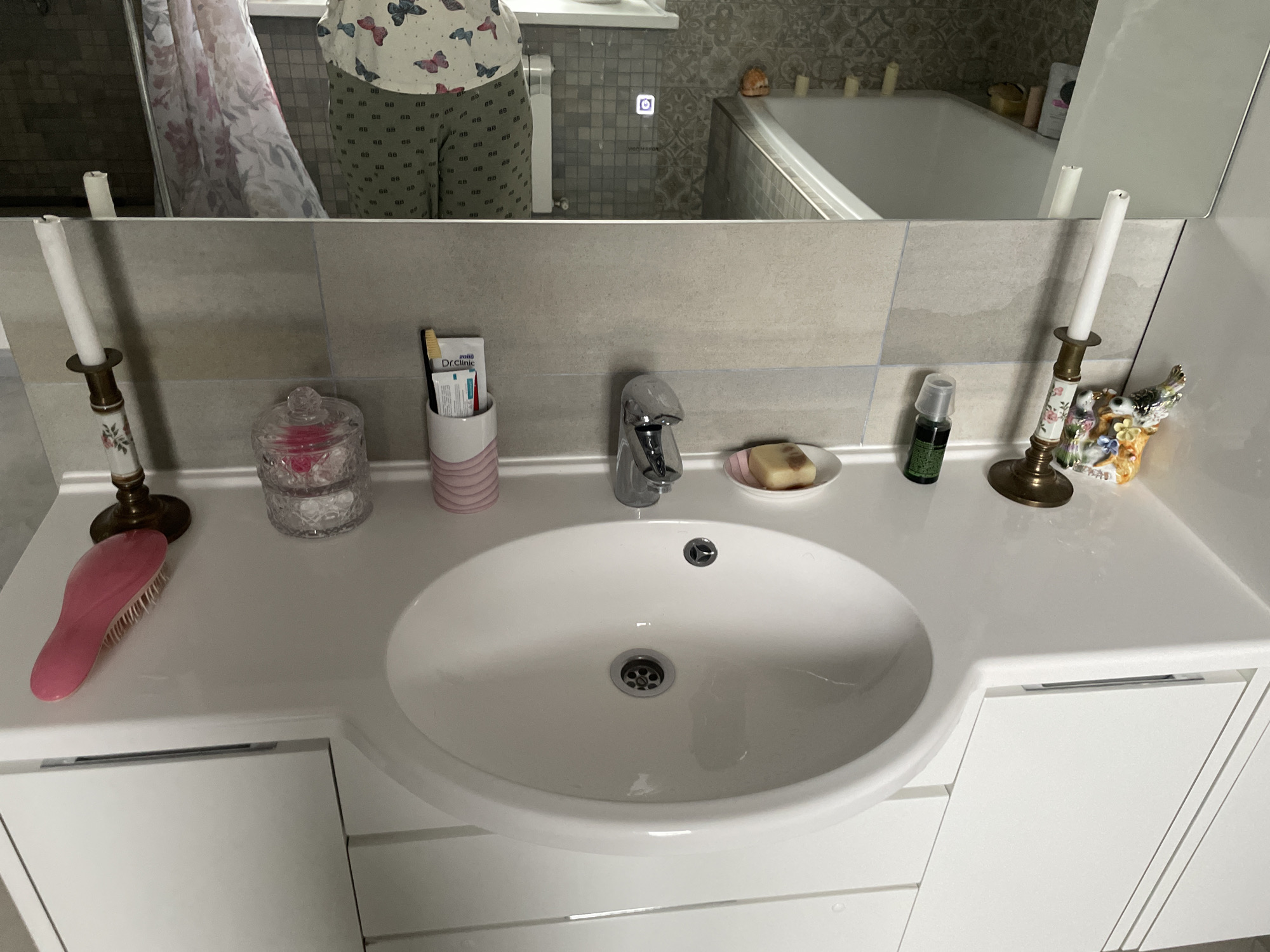 Here is a forensic description of the image:
**Scene:** The image appears to be a close-up shot of a bathroom vanity. The vanity is white and features a white, oval-shaped sink. A chrome faucet is mounted on the counter behind the sink.
**Objects:**
* **Sink area:** A pink hairbrush, a decorative glass jar (presumably holding cotton balls or similar items), a toothbrush holder with toothbrushes, a bar of soap on a small white dish, and a small bottle of dark liquid (possibly a facial serum or essential oil) are arranged on the counter.
* **Decorative items:** Two tall candlesticks with white candles are positioned at either end of the counter. A small, decorative porcelain figurine is placed near one of the candlesticks.
* **Mirror reflection:** The reflection in the bathroom mirror shows a person wearing pajamas and standing in front of the sink. It also shows a bathtub and window in the background.
**Environment:**
* The background consists of gray tiles above the vanity.
**Location:**
* Based on the description, this could be a bathroom in a home or apartment located in Лесной Пейзаж Кп (шарапово), Russia.