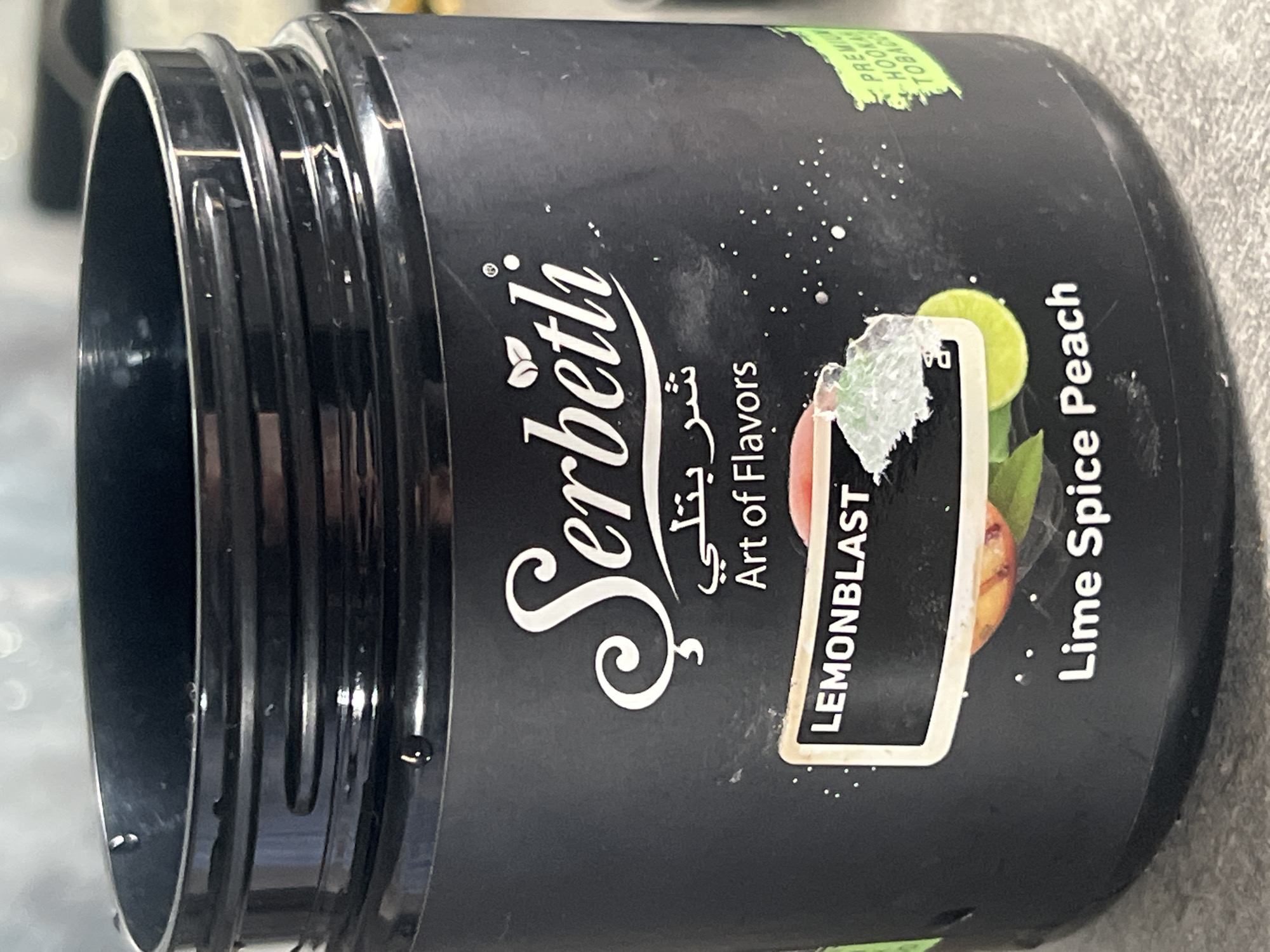The image shows a black container of Serbetli brand hookah tobacco. The flavor is "Lemonblast Lime Spice Peach." The label also says "Art of Flavors" in English and Arabic. The container is likely sitting on a gray surface. No information in the image indicates a location in Yerevan, Armenia.