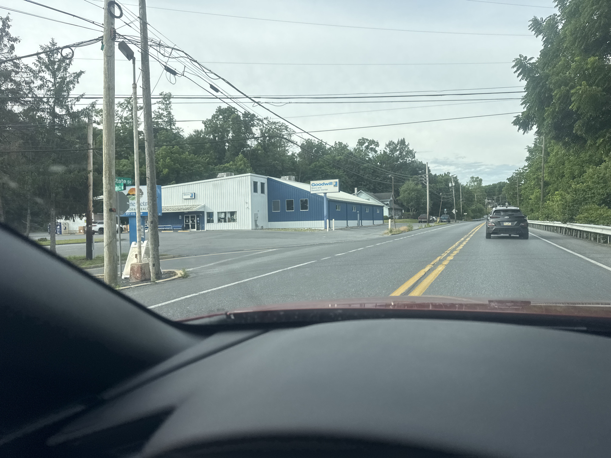 This media file captures a view from inside a vehicle, looking out at a suburban street scene. The main subjects are a blue and white Goodwill store with a blue sign that reads "Goodwill Store and Donation Center," and a road with a black SUV in the distance. A metal guardrail lines the right side of the road, and a line of trees is visible on the right, with more trees and utility poles on the left. The sky is overcast, suggesting a cloudy day.
The scene appears to be set during daylight hours, likely mid-morning or afternoon. There are no people clearly visible in the image, only the car and the building. The activity taking place is simply the view of a commercial establishment and a road, suggesting a travel or drive-by perspective.
Notable details include the prominent Goodwill signage, the two utility poles on the left side of the road with a complex network of wires overhead, and a green street sign that appears to say "STATE ST" followed by more text that is unreadable. The building has multiple windows and a blue awning. The road has double yellow lines indicating it is a two-way street. The perspective from inside the car shows the car's dashboard and windshield, framing the external view. There is no discernible emotional tone conveyed, as it's a straightforward depiction of a roadside scene.