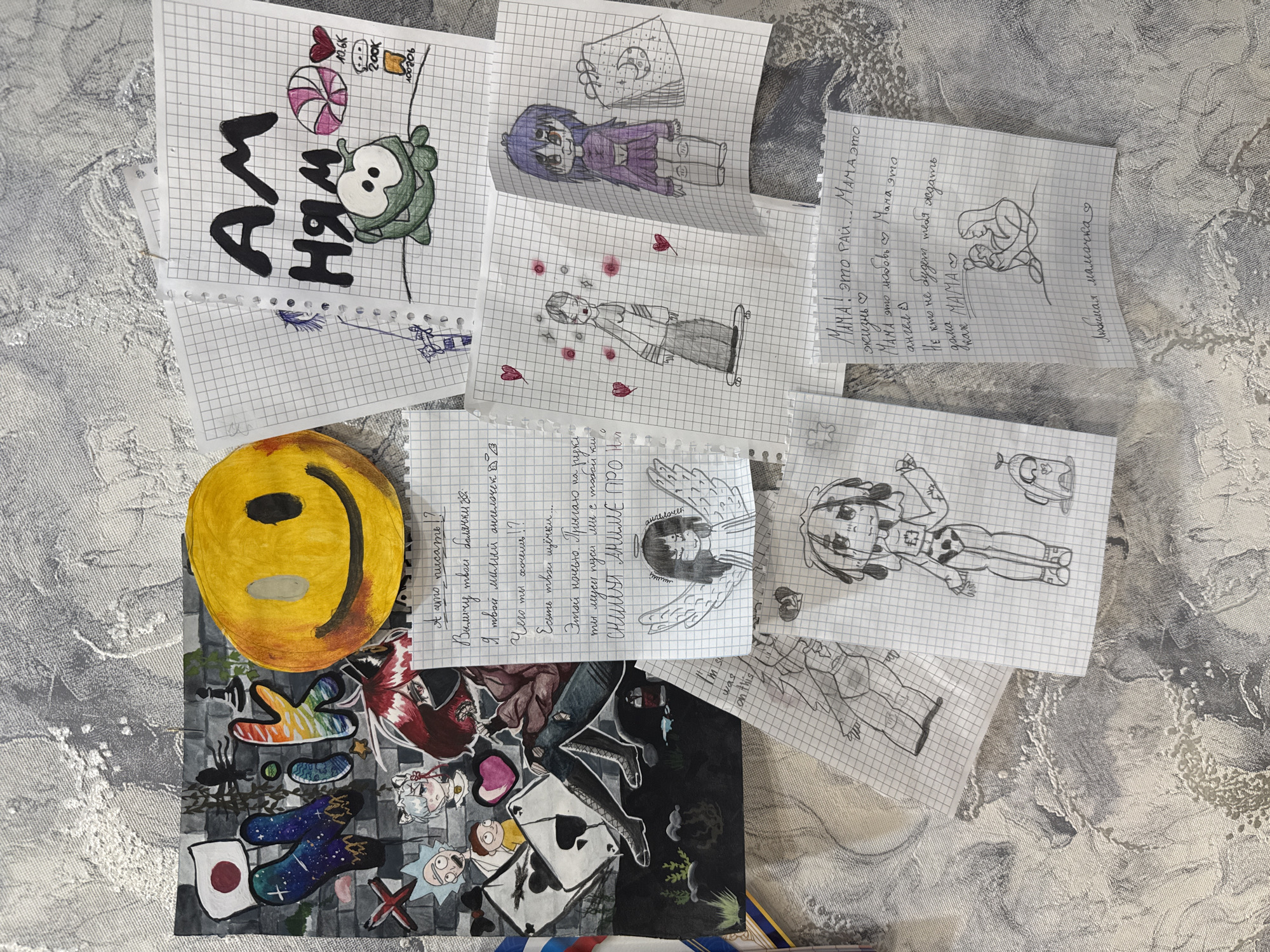 The image shows a collection of drawings and artwork scattered on a patterned surface. The drawings, done on graph paper, include anime-style characters, a cartoon monster with a lollipop, and a depiction of a mother and child. Some pages contain text in Russian. A painted smiley face and a collage featuring various pop culture references are also visible. It cannot be determined from the image whether the location is in the city of Rusanovo, Russia.