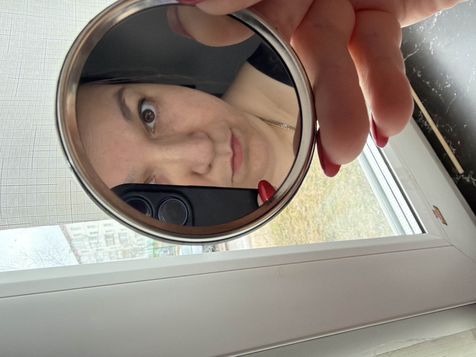 A close-up, eye-level photograph captures a woman's reflection in a handheld circular mirror. The woman has dark hair, a light complexion, and is wearing a necklace. Her right eye is wide open, giving a surprised or playful expression, while her mouth is slightly smirking. A hand with red polished nails holds the mirror, with a black smartphone, featuring a prominent camera module, also visible in the reflection.
The setting is indoors, near a window. To the left, a light-colored, patterned curtain or blind is visible. To the right, a dark, possibly marbled, surface is present. The white-framed window reveals an outdoor scene with bare tree branches and distant multi-story buildings, suggesting an urban environment in Chaikovsky, Russia. The natural light indicates it is daytime. There is no visible text in the image.
