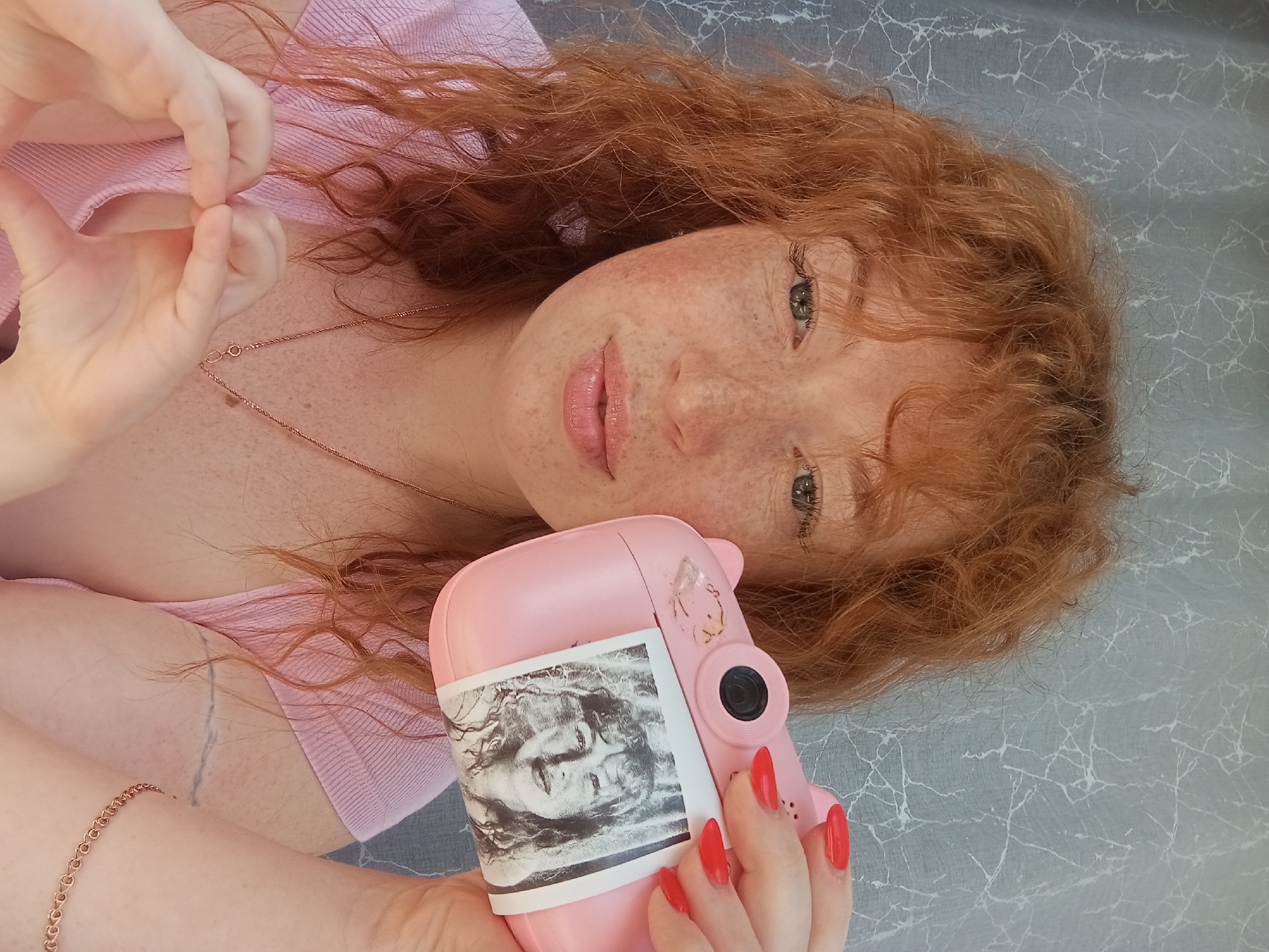 A woman with red hair and freckles, holding a pink camera with a black and white photo attached to it, lies on a grey patterned surface. She makes a heart shape with her fingers. She wears a pink shirt and a gold necklace. Her fingernails are painted red. The scene is indoors and there is no indication of the location being in the city of Yurgamysh, Russia.