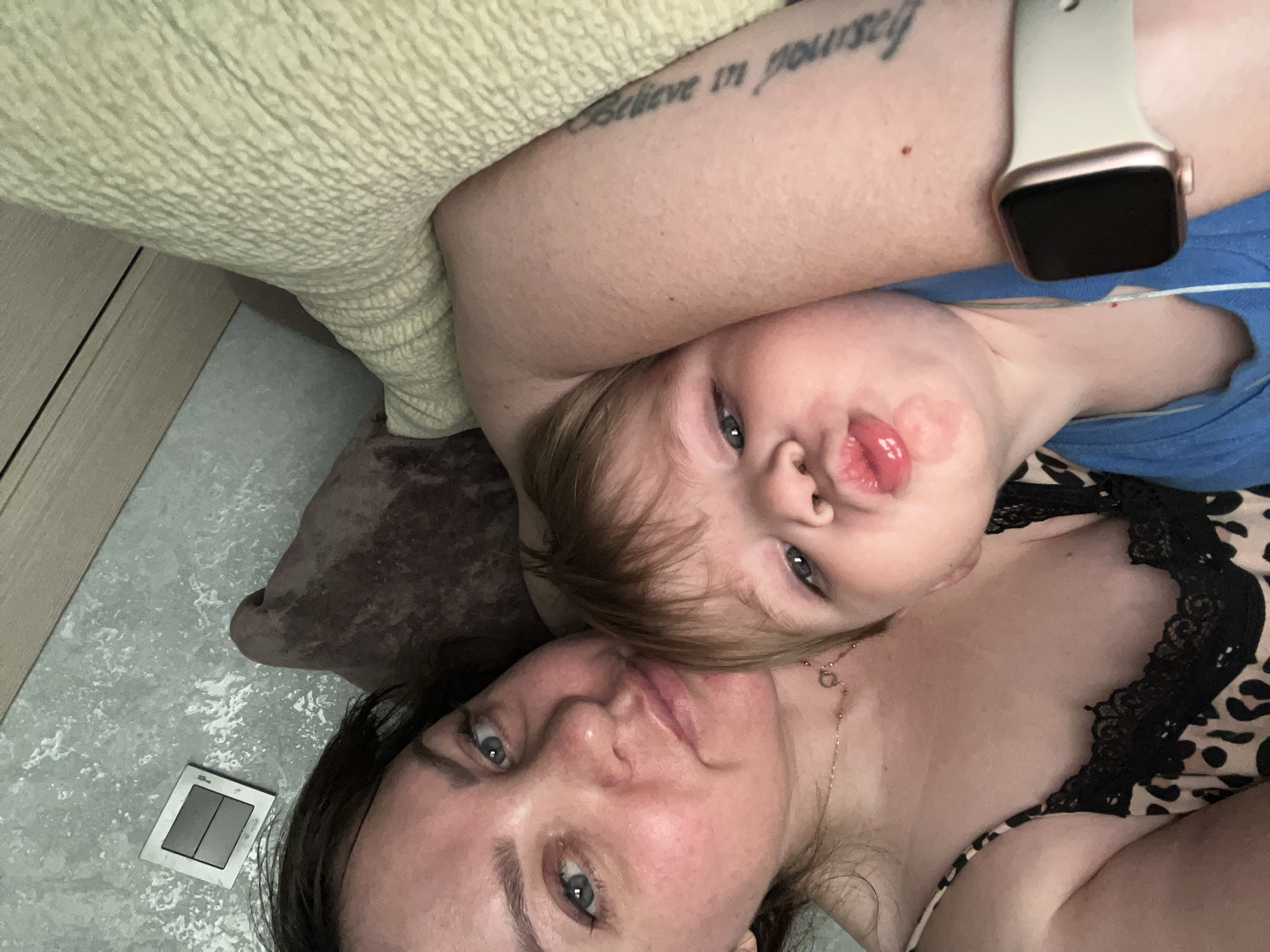 A woman and a young child are lying down, possibly in a bed or on a couch, in a room. The woman is wearing a leopard-print top with black lace trim and a thin necklace. She has dark hair and blue eyes. The child, with light brown hair and blue eyes, is nestled against the woman and making a puckering expression. The child is wearing a blue shirt. On the woman's left wrist, a smartwatch with a light-colored band is visible. A tattoo can be seen on the woman's arm. In the background, a textured wall is visible with a light switch. The overall lighting suggests an indoor setting during the day or evening. The image was taken in Noviy, Russia.