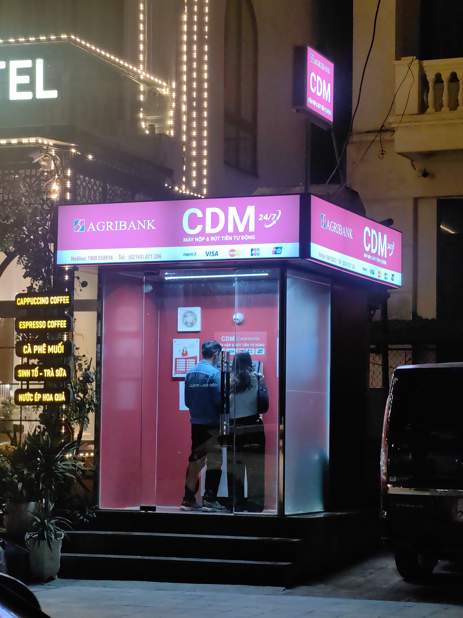 An Agribank Cash Deposit Machine (CDM) booth is centrally positioned on an urban street in Sa Pa, Vietnam, at night. The illuminated, glass-enclosed booth features a prominent pink Agribank CDM 24/7 sign overhead, displaying a hotline, telephone number, and accepted card types including Napas, Visa, and Mastercard. Two individuals are inside the booth; a male in a blue jacket stands to the left, and a female in a light jacket stands to the right, both facing individual ATM machines.
To the immediate left of the booth, a vertical stack of five yellow illuminated signs advertises beverages: "CAPPUCCINO COFFEE," "ESPRESSO COFFEE," "CÀ PHÊ MUỐI" (Salted Coffee), "SINH TỐ - TRÀ SỮA" (Smoothie - Milk Tea), and "NƯỚC ÉP HOA QUẢ" (Fruit Juice). Further left, a building adorned with strings of warm white LED lights displays a partial "TEL" sign, indicative of a hotel. Potted plants are arranged near the base of the beverage signs and the hotel entrance.
On the right side of the scene, a darker building with a white classical-style balcony railing is visible. A dark-colored vehicle, likely an SUV or van, is partially parked on the far right, facing away from the viewer. Steps lead up from the pavement to the entrance of the ATM booth. The scene is brightly lit by the various signs, contrasting with the dark background of the night.