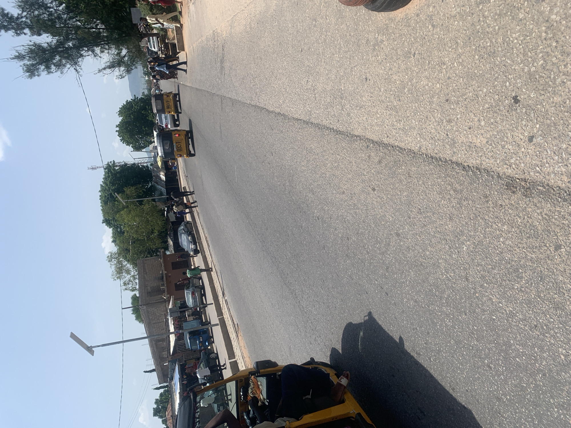 The image depicts a street scene in Jalingo, Nigeria, under a clear blue sky, suggesting it is daytime. The perspective is from a low angle, looking down the road, which is paved and appears to be in moderate condition with a visible centerline.
The scene is populated with several three-wheeled vehicles, commonly known as "keke-napep" or auto-rickshaws, which are a popular mode of transportation in Nigeria. Some of these vehicles are yellow, a common color for them, and others appear to be white. People are visible interacting with the vehicles and along the sides of the road, indicating a busy street. Some individuals are standing, while others might be passengers or drivers.
Along one side of the road, there are buildings, some appearing to be made of brick, and others with corrugated metal roofing. There are also some trees providing shade, and a lamppost with a solar panel is visible, suggesting a mix of development and natural elements.
The activity appears to be the normal hustle and bustle of a Nigerian street, with vehicles and pedestrians moving along. There are no overt signs of distress or unusual events. The overall impression is one of a typical day in a populated urban or semi-urban area in Nigeria, characterized by local transportation and daily life. The image is slightly tilted, giving a dynamic feel to the scene.