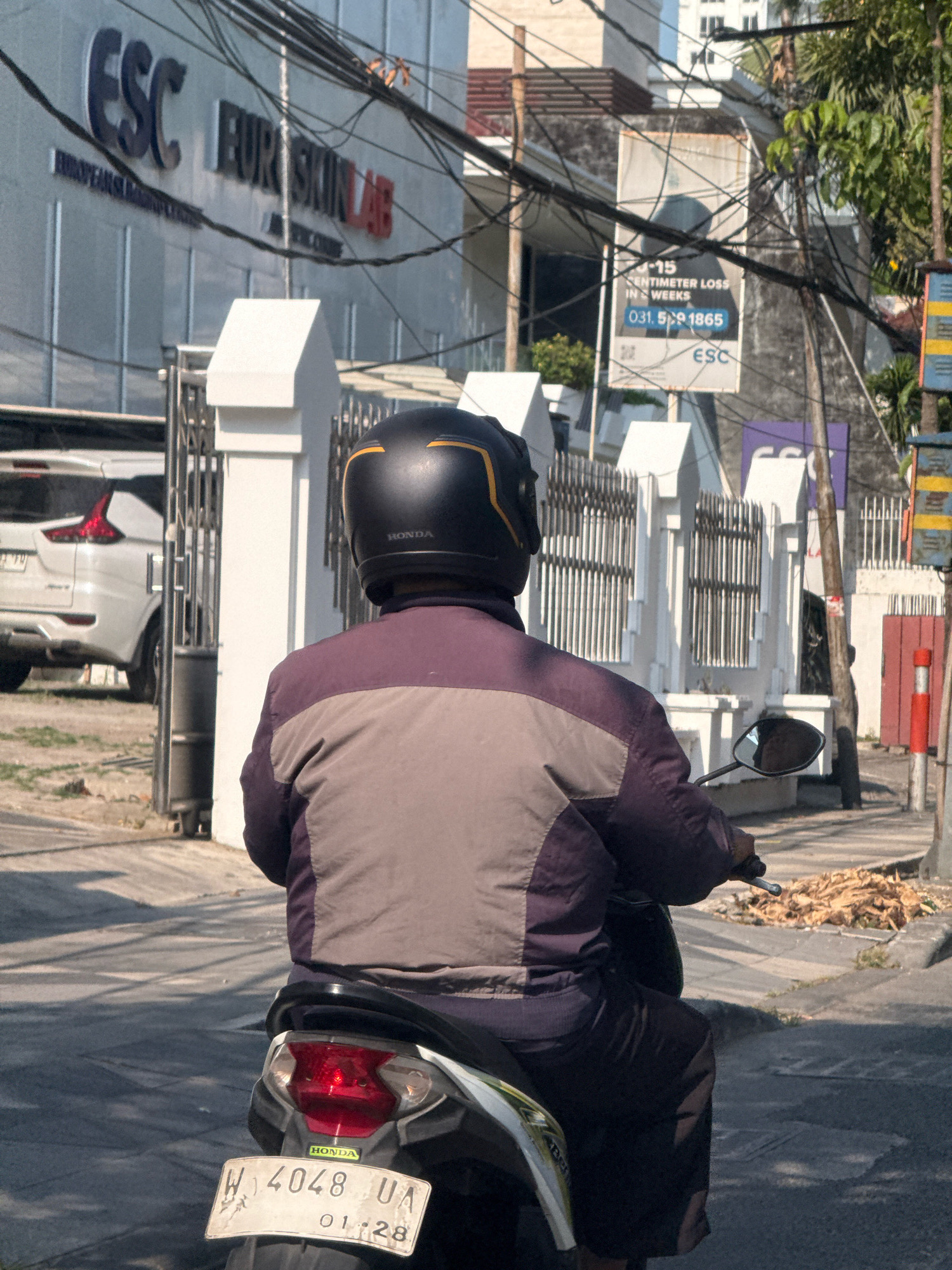 An over-the-shoulder view shows a person on a motorcycle from behind, facing away from the camera and riding down a street in Surabaya, Indonesia. The rider is wearing a black helmet with a yellow stripe and a jacket with maroon and grey panels. The motorcycle has a license plate that reads "W 4048 UA 01-28". In the background, a white building with "ESC" and "EUROPEAN SKIN LAB" signage is visible, along with a fence and a white SUV. A sign on a pole in the background advertises a "15 CENTIMETER LOSS IN 4 WEEKS" with a phone number "031-5991865". The scene is bathed in sunlight, casting shadows on the road.