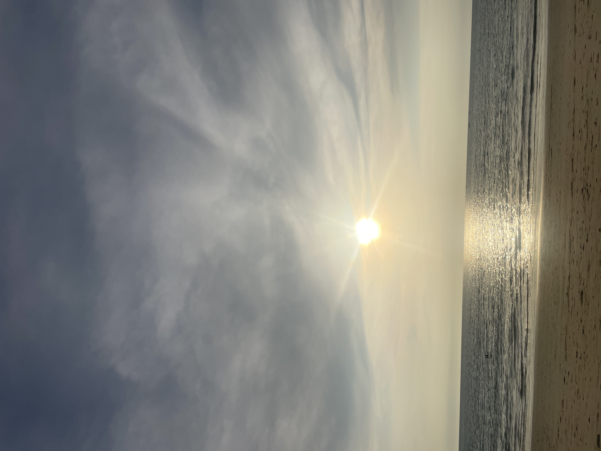 The image captures a beach scene in Choeng Thale, Thailand. The sun is high in the sky, partially obscured by clouds, casting a bright glare. The ocean is visible, with sunlight reflecting off the water's surface. The shoreline is partially visible on the right side of the frame. No people are distinctly visible.