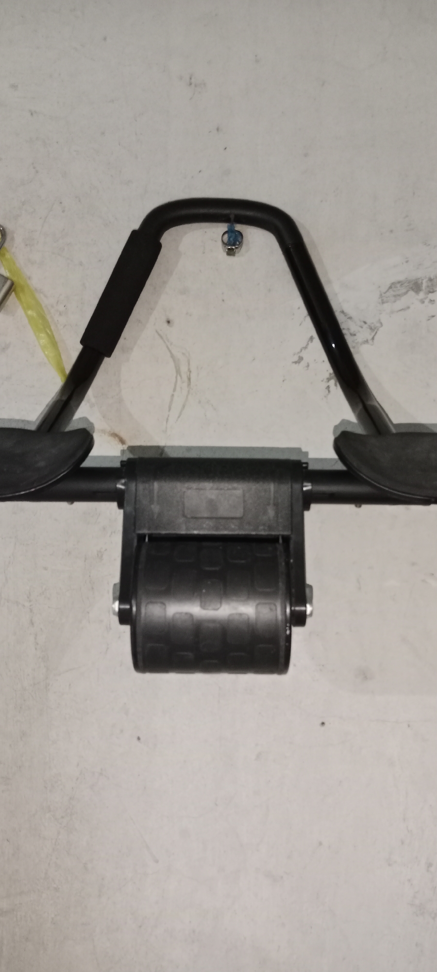A close-up, head-on shot shows the front of a black, metal exercise roller with black padded armrests attached to the curved top bar. The roller has a textured surface and is mounted on a black bar that extends horizontally. The roller and bar assembly is suspended against a pale, textured wall that appears to be made of concrete or a similar material. A blue cord is tied to a bolt on the upper bar. The scene is lit from the front, casting minimal shadows. There are no people present, and no activities are taking place. The overall setting is that of an indoor space, possibly a gym or a storage area, with no indication of its specific location in Subang, Indonesia.