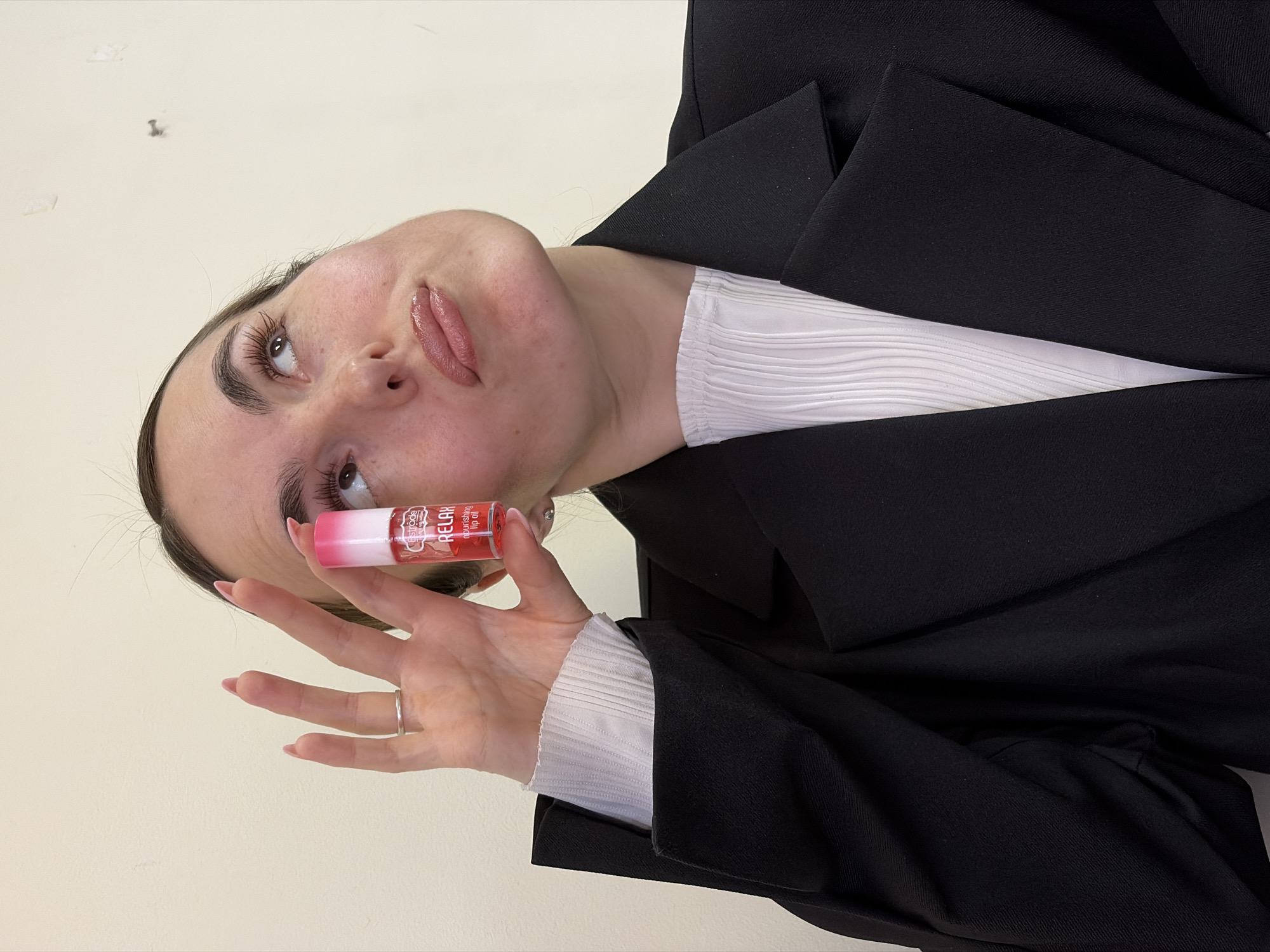 A young woman with fair skin, dark hair, and visible freckles is holding a tube of lip gloss in her right hand. The lip gloss has a pink and white gradient top and the words "RELAX nourishing lip oil" visible on the label. She is wearing a silver ring on her right ring finger. She has light pink lipstick on. She is wearing a white, textured top and a black blazer. The background is a plain, light beige wall. The photograph is taken from a low angle and rotated 90 degrees counter-clockwise. There is no information indicating the location is Izhevsk, Russia.