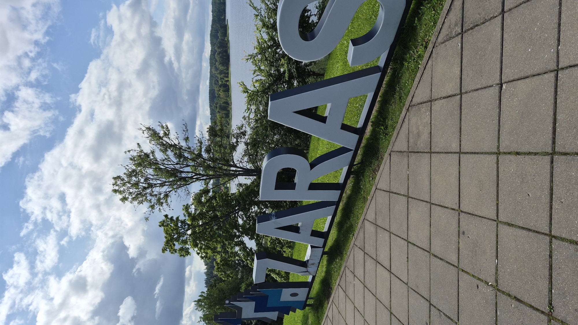 The image shows a close-up of large, three-dimensional, white letters spelling "ZARASAI" with a dark blue outline. The letters are situated on a grassy slope in front of a body of water and trees. To the left of the letters, there's a stylized graphic element in shades of blue and grey, also appearing to be part of the signage. The foreground is a paved area made of square concrete tiles with some grass growing between them. In the background, a calm lake or river is visible, bordered by a dense line of green trees and a distant, undulating landscape under a sky with scattered white clouds. The setting appears to be an outdoor public space, likely a park or waterfront promenade, in the city of Zarasai, Lithuania. There are no people visible in the image.