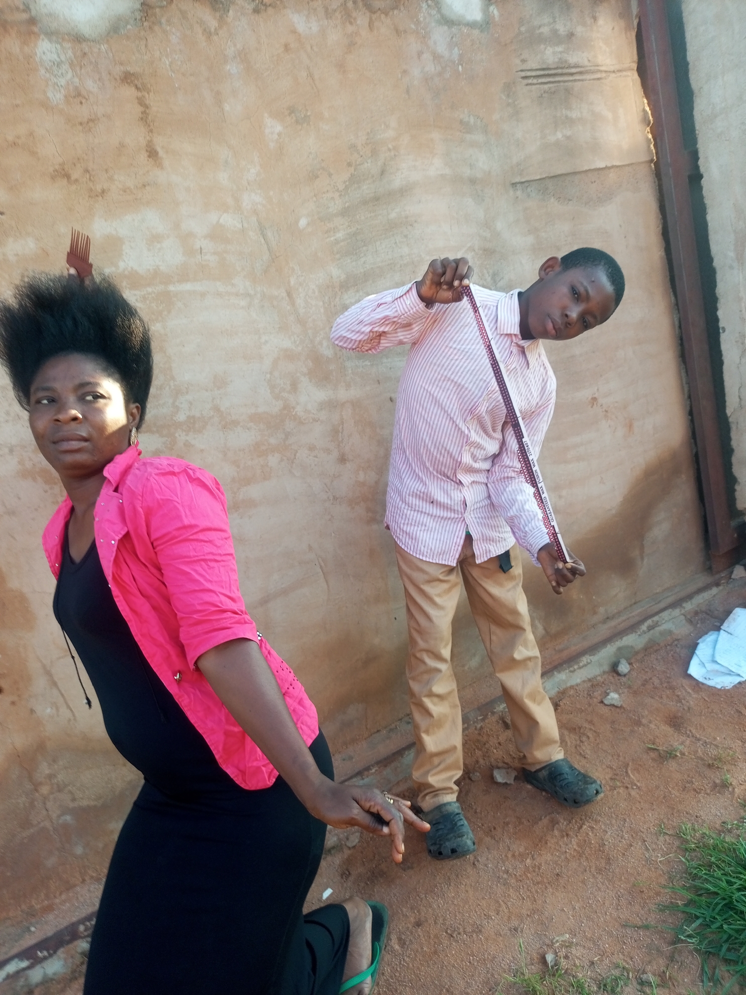 A full shot shows two individuals posed in front of a stucco wall. On the left, a woman with voluminous dark hair stands to the left, facing towards the camera but looking away. She is wearing a bright pink blazer over a black dress, and a comb is stuck in her hair. On the right, a young boy in a pink and white striped shirt and tan pants stands and holds a measuring tape. His left hand is near the top of the tape, and his right hand is near the bottom, extended towards the ground. The ground beneath them is a reddish-brown dirt mixed with small rocks and sparse green grass. The wall behind them is textured and weathered, with some visible cracks and a metal beam running vertically on the right side of the frame. The lighting suggests it is daytime. The scene appears to be an outdoor setting, potentially in a less developed area.
