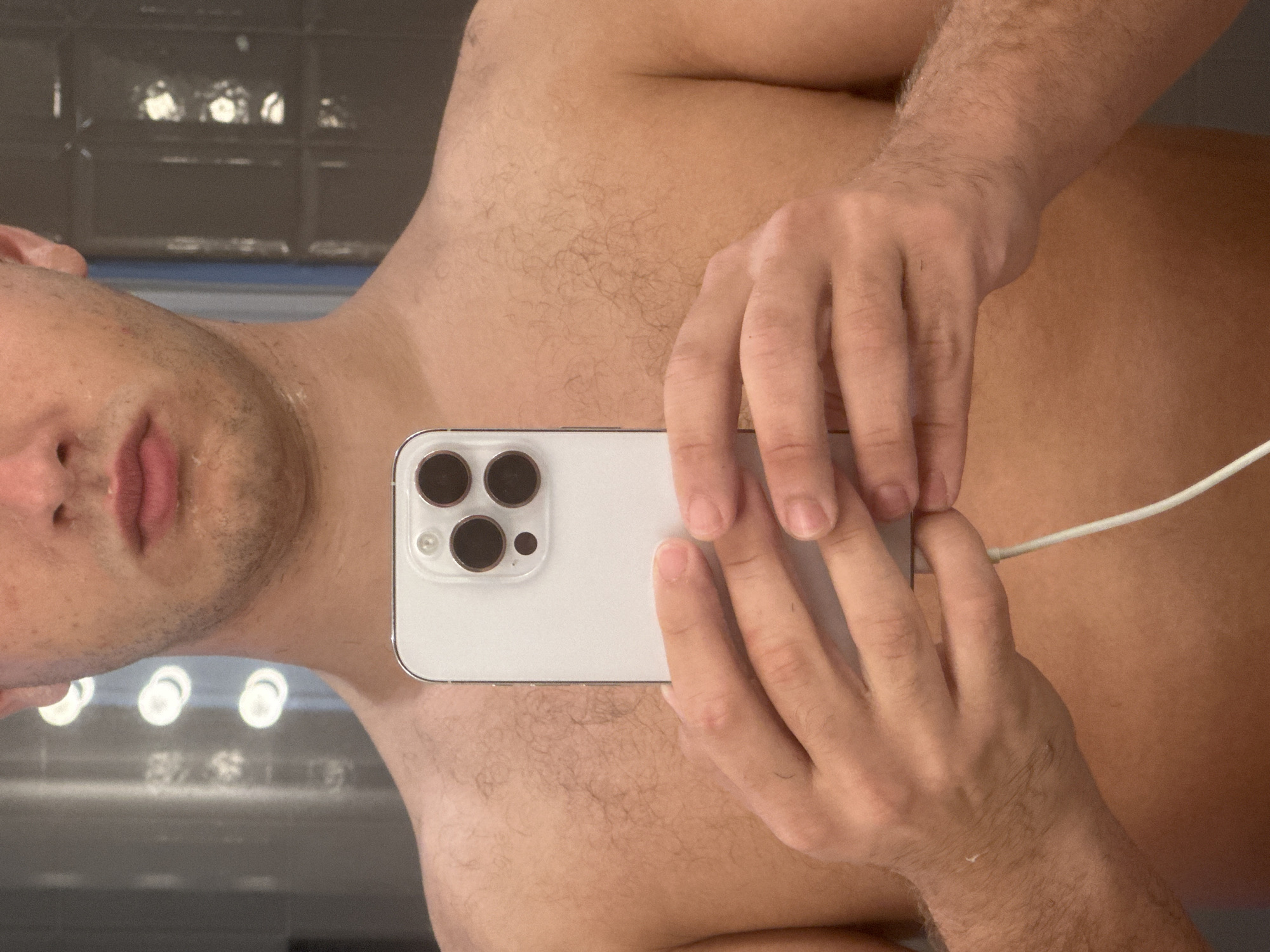 A selfie photograph depicts a shirtless male holding a white iPhone, possibly an iPhone 13 Pro or similar model, in front of his body. The phone is plugged into a charger. The background reveals a gray tiled wall and a row of lights, potentially suggesting a bathroom setting. The man's face is visible, though partially obscured by the phone. There is no information to indicate the photograph was taken in Milan, Italy.