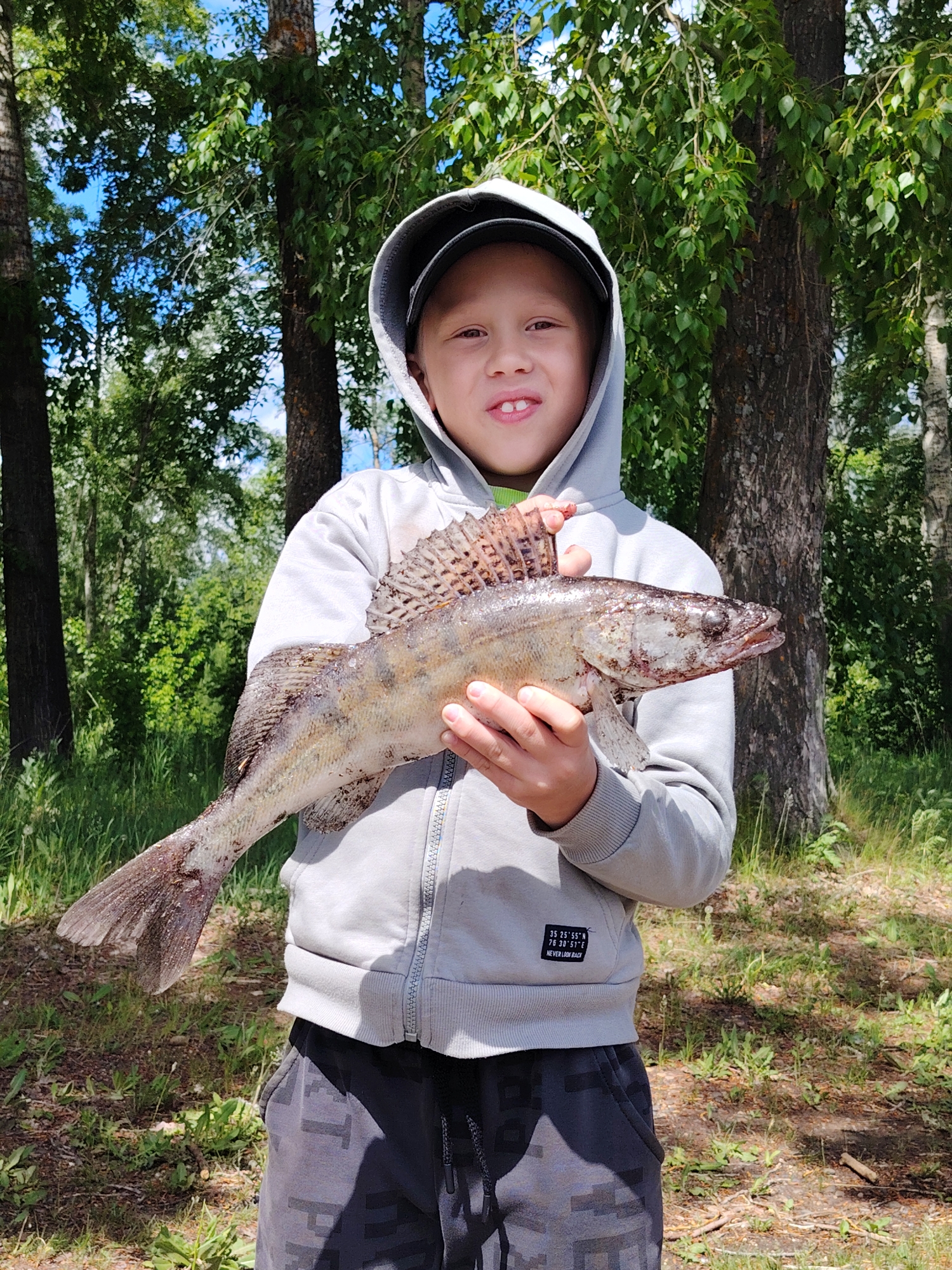 A young boy, approximately 6-8 years old, stands outdoors, holding a large fish. He is wearing a gray hooded sweatshirt, dark shorts, and a baseball cap under the hood. The fish is held horizontally in front of him, with the boy's hands supporting its body. The fish is speckled and appears to be a zander. The background includes trees and foliage, suggesting a park or wooded area. Based on the coordinates printed on the sweatshirt (35° 25' 55" N, 76° 30' 51" E), it cannot be verified that it is in Chaikovsky, Russia.