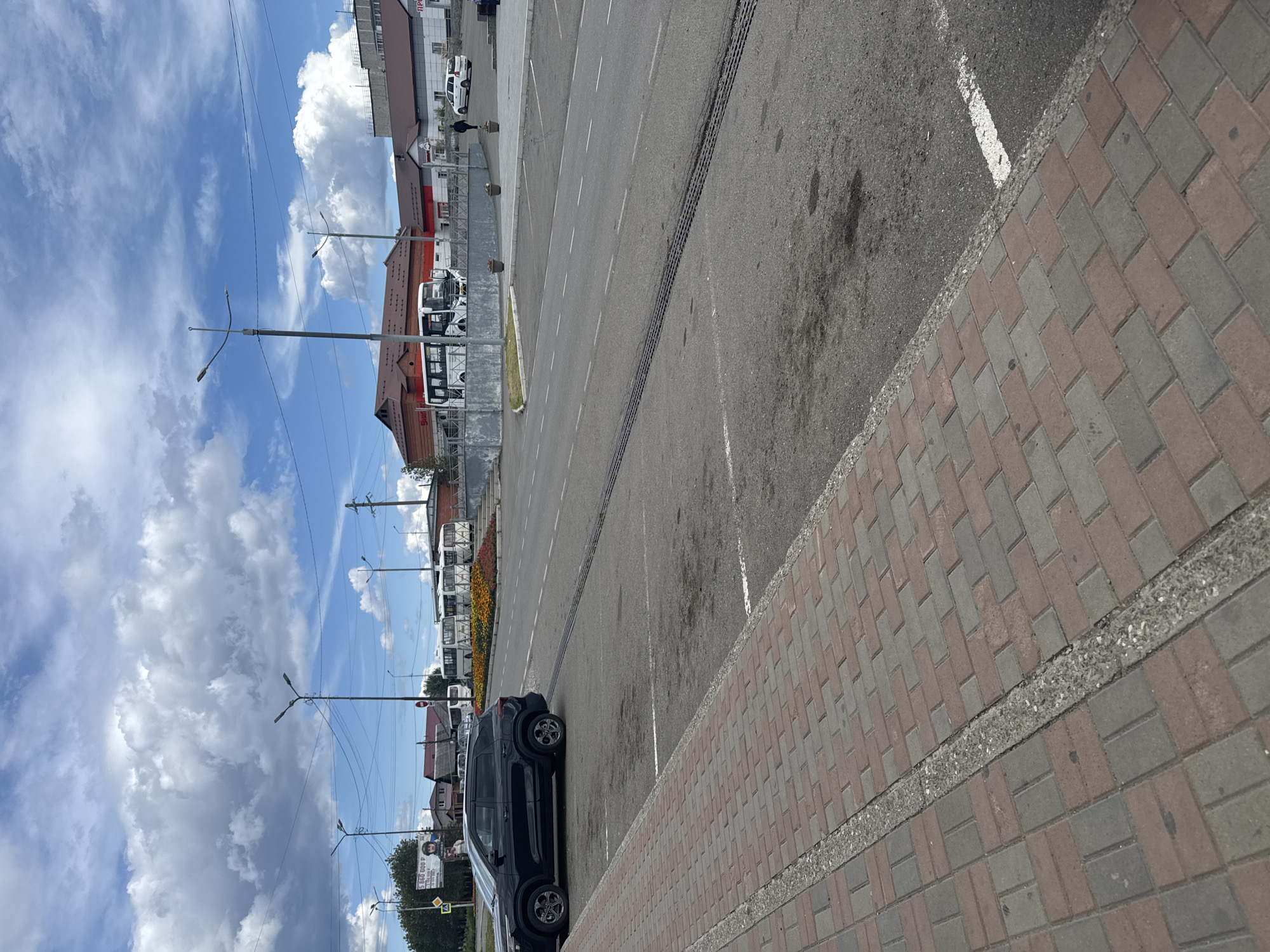 A wide shot of a street and parking area on a cloudy day. In the foreground, a red and gray brick sidewalk runs along the left side of the image. Beyond the sidewalk is a paved parking lot with white parking lines. A black SUV is parked in the lot. In the background, a street curves to the right, with several white buses parked in a lot to the left. Buildings are visible behind the buses. Streetlights and power lines are strung across the sky.