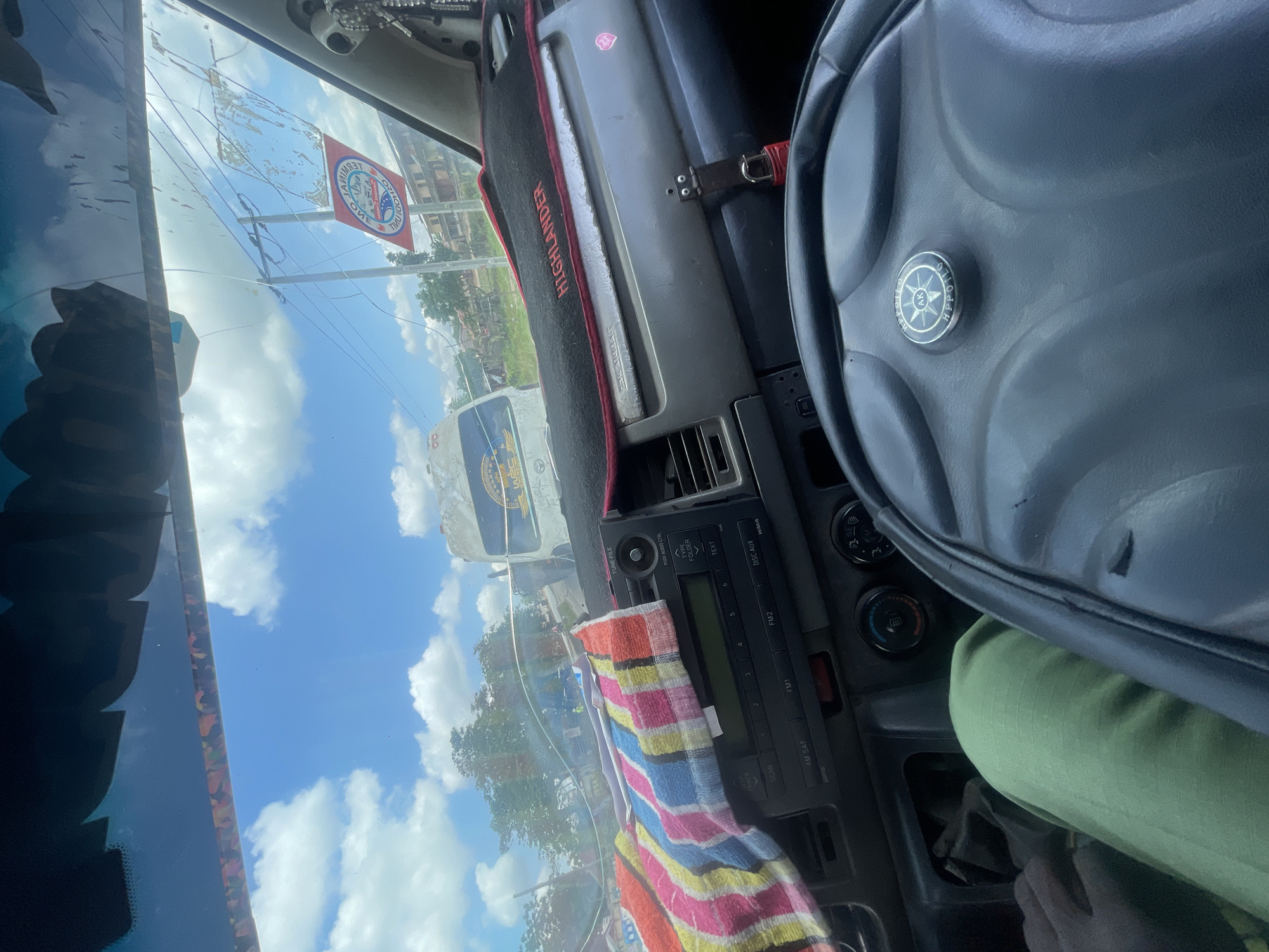 The image is taken from the interior of a vehicle, looking forward through the windshield. The dashboard is visible, with a colorful striped cloth draped over the center console, partially obscuring a car stereo. The steering wheel is in the foreground, to the right. Outside the vehicle, a bright blue sky with scattered white clouds is visible. Several vehicles, including a white bus with signage, are seen on a road, suggesting a city or town environment. Trees and buildings are present in the background. A red and white circular sticker with text is attached to the windshield. The lighting suggests it is daytime. Based on the context provided, the location is the city of Ikire, Nigeria.