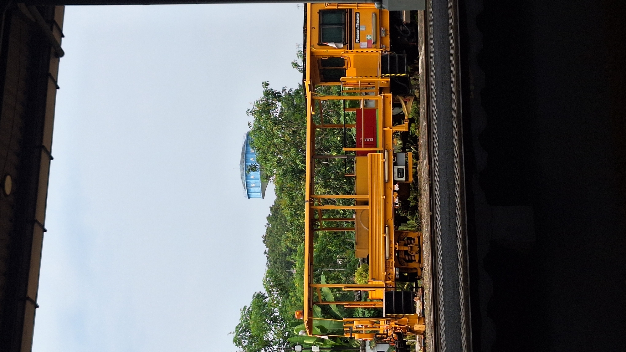 The image shows a yellow maintenance vehicle on a railway track, framed by the structure of a building, possibly a warehouse or industrial shed, with the sky visible in the distance. The vehicle has a complex frame, and is parked on tracks with green vegetation visible nearby. In the background, beyond the railway and vegetation, a water tower with "KARAWANG" inscribed is noticeable. The scene is likely in or around Karawang, Indonesia, due to the prominence of the city's name on the water tower.