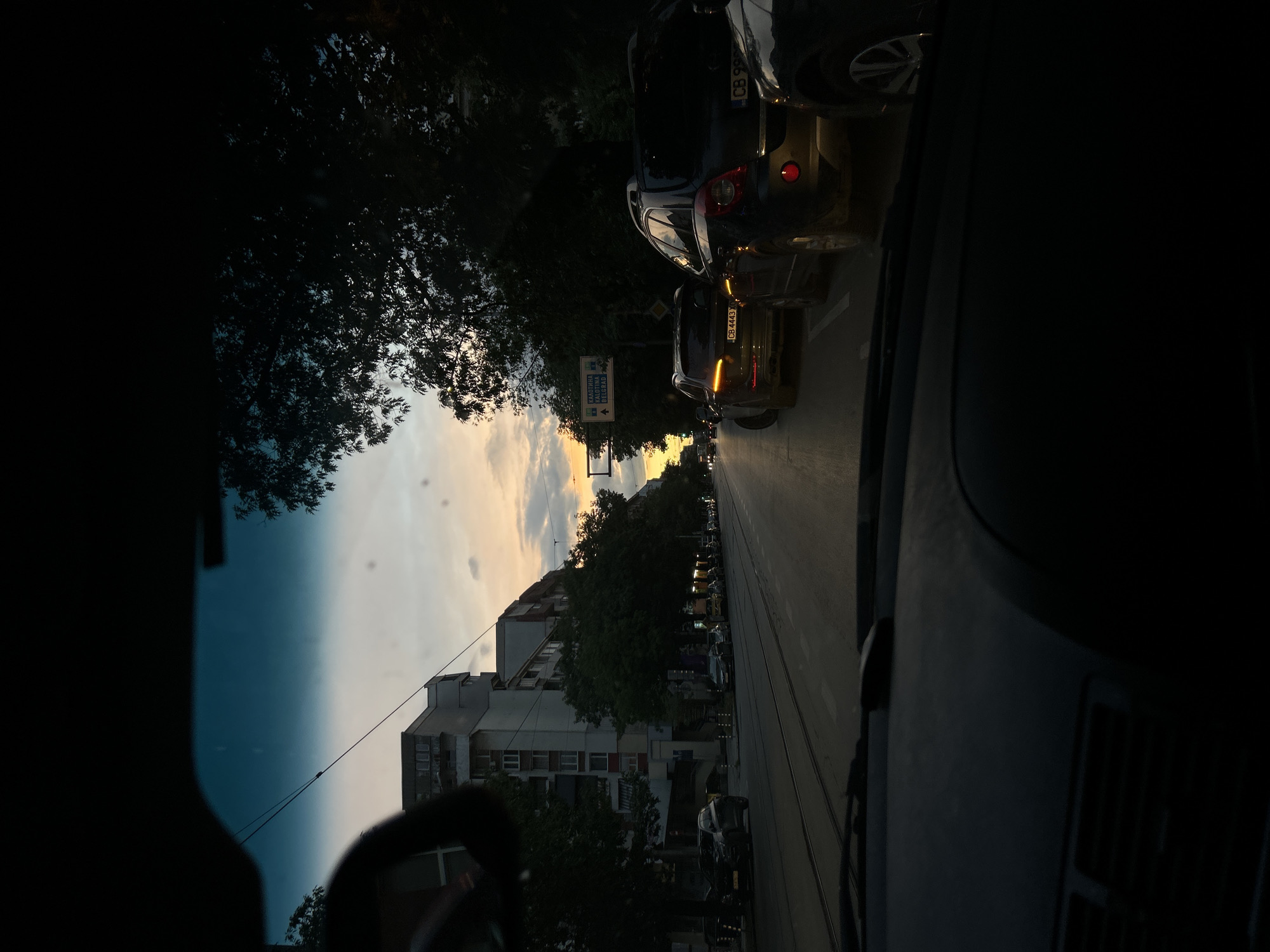 The image depicts a street scene in a city at sunset, viewed from the interior of a vehicle. In the foreground, the dashboard and windshield of the car are visible, framing the external view. The road ahead is lined with parked cars and traffic, suggesting congestion. On the left side of the street, apartment buildings and trees line the sidewalk. On the right, dense foliage obscures the view of the buildings behind it. The sky displays hues of orange, yellow, and gray, characteristic of a sunset. Overhead, power lines crisscross the sky. A street sign with Bulgarian text is visible on the right side of the road, indicating directions to Kalotina and Belgrade. The overall impression is of a typical urban environment during rush hour, with the setting sun casting a warm glow over the scene.
