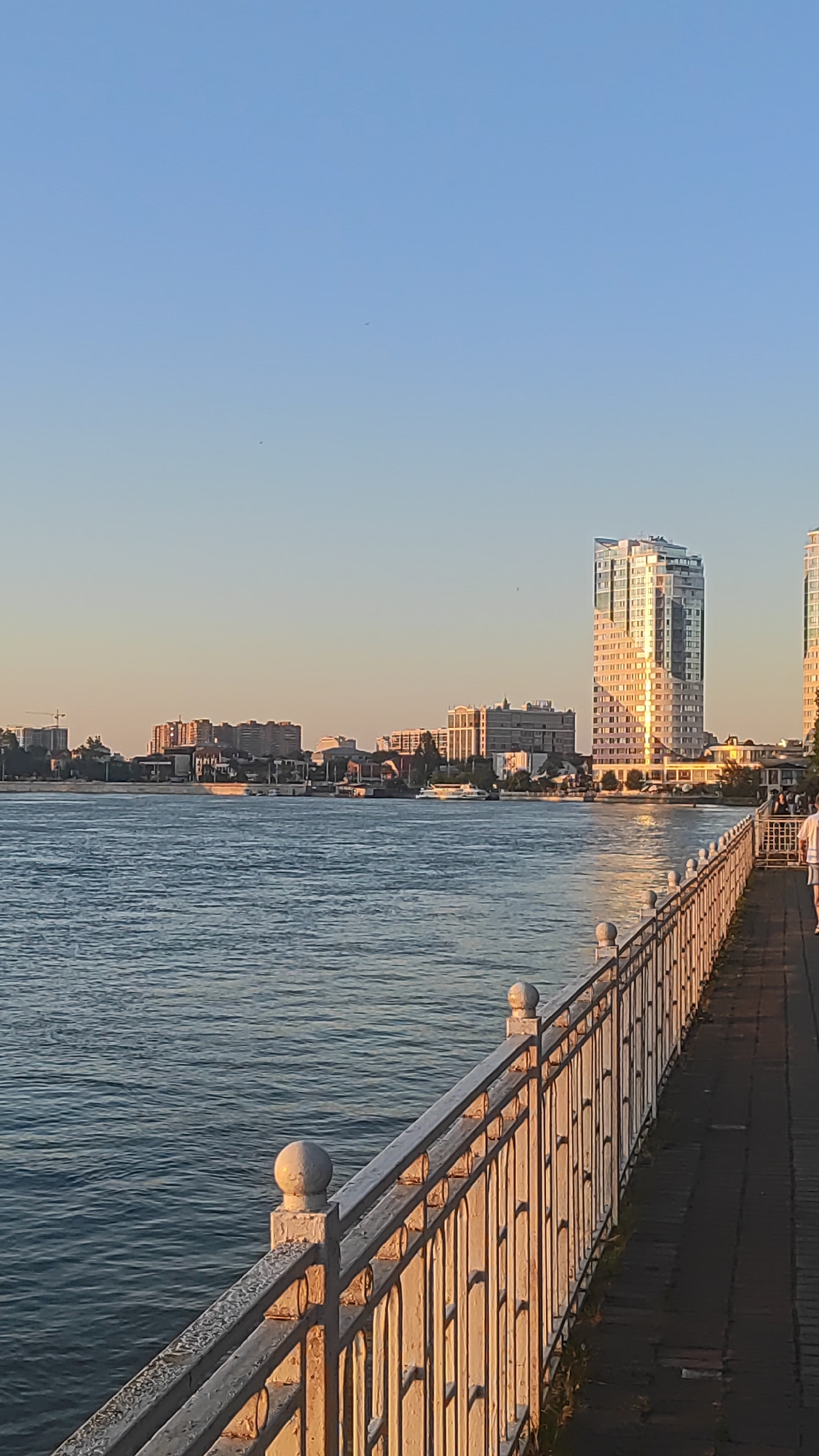 This media file depicts a scenic waterfront promenade with a city skyline in the background. The scene is bathed in the warm, golden light of either sunrise or sunset, casting long shadows and highlighting the architectural features of the buildings. The sky is clear and a soft blue, indicating pleasant weather.
The foreground features a paved walkway alongside a decorative metal railing that separates it from a wide body of water. The railing is white with spherical finials on its posts and shows some signs of age and weathering, adding character to the scene. The water appears calm with gentle ripples reflecting the light.
In the middle ground, across the water, a city skyline is visible. It consists of various buildings, including several apartment complexes and office buildings. A prominent, modern high-rise building with a reflective facade stands out, catching the sunlight. Several boats, including what appears to be a white passenger ferry, are on the water. The distant shore is lined with trees and smaller buildings, suggesting a developed urban area.
In the background, the clear sky transitions to a lighter hue near the horizon, characteristic of dawn or dusk. There are no discernible text elements visible in the image. The overall mood of the image is peaceful and serene, evoking a sense of calm and urban beauty. The presence of a few people walking on the promenade suggests it's a public space enjoyed by residents and visitors. The location context of Krasnodar, Russia, places this scene along one of its waterfronts, likely a popular spot for recreation and enjoying the cityscape.