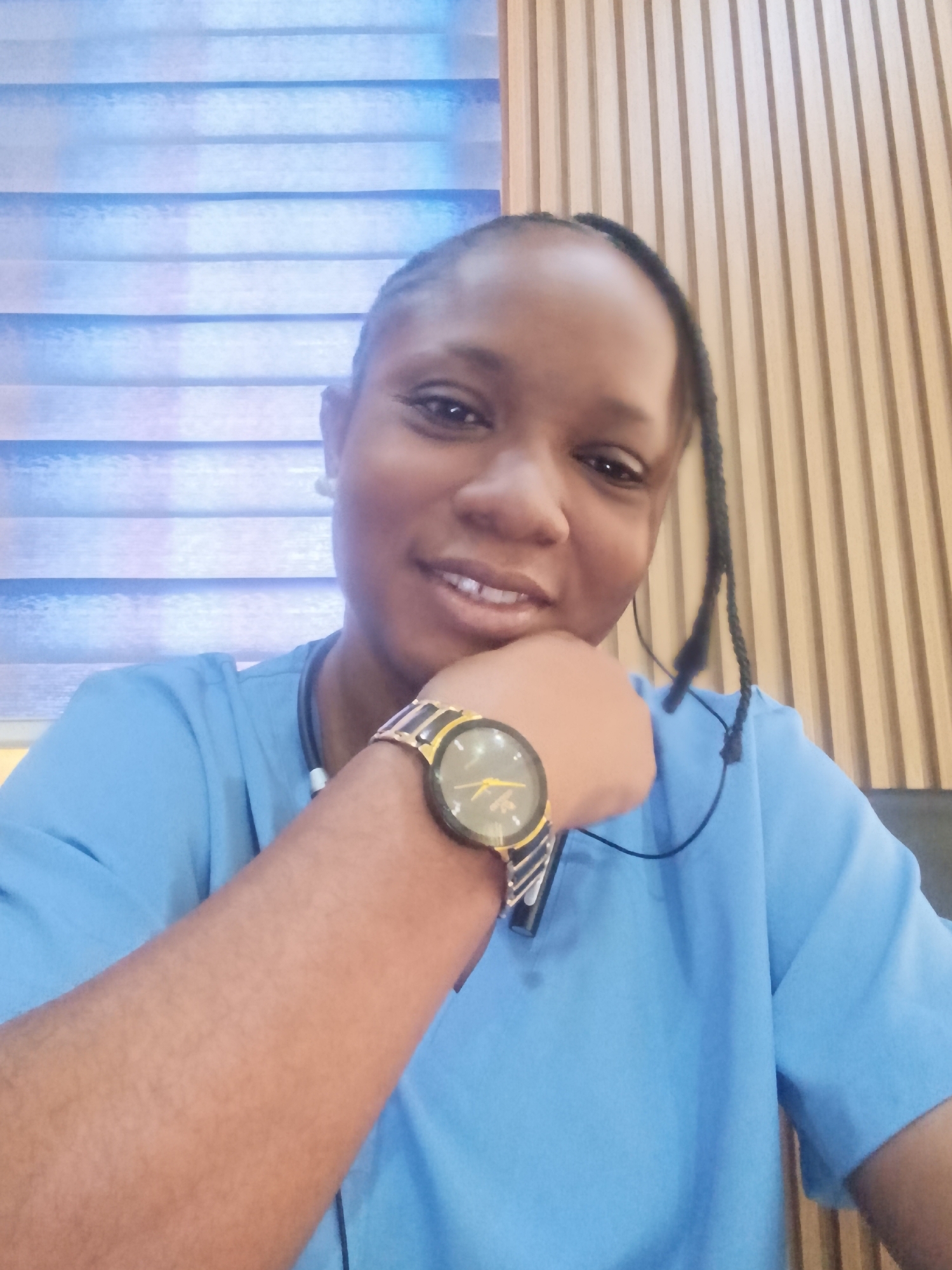 A medium shot depicts a dark-skinned woman wearing a light blue shirt. She has a watch on her left wrist and her left hand is supporting her chin. She has braids. A stethoscope hangs around her neck. The background consists of blue and white window blinds on the left and vertical wooden slats on the right. The location is presumed to be in Gwarinpa, Nigeria, but there is no specific detail in the image to confirm this.