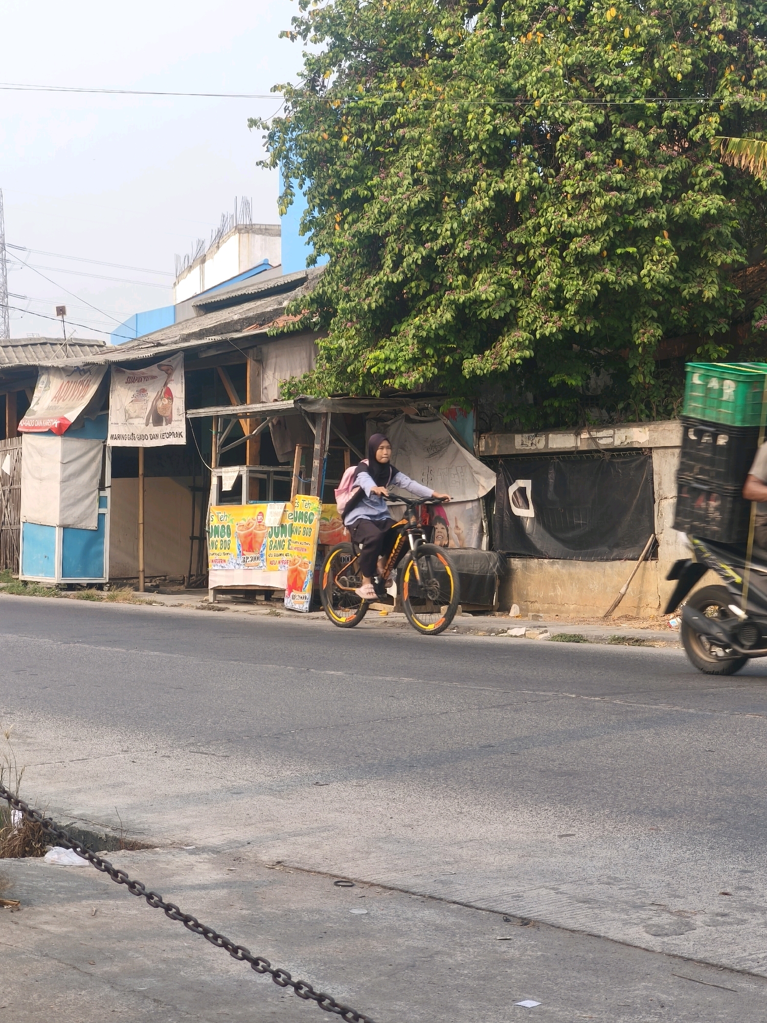 The image depicts a street scene in the city of Bekasi, Indonesia, with a focus on the roadside businesses and activity. A young woman wearing a hijab is riding a bicycle on the asphalt road. She is wearing a blue long-sleeved shirt and dark pants, and a pink backpack is strapped to her back. The bicycle has an orange frame and yellow-accented wheels. To her right, a motorbike is visible, carrying stacked green plastic crates in the back.
Along the roadside, there is a row of modest structures that appear to be small shops or food stalls. One stall displays bright posters advertising "Teh Jumbo Bob" with images of iced drinks. Other banners are hung above the stalls, one with text that includes "WARUNG GADO GADO DAN KETOPRAK." The buildings have corrugated metal roofs and open fronts. Behind these stalls, a large tree with abundant green foliage and some yellowing leaves dominates the upper right portion of the image. The sky is overcast with a few thin clouds. In the foreground, a heavy metal chain lies on the ground along the edge of the road. The overall environment suggests an urban setting with informal commercial establishments.
