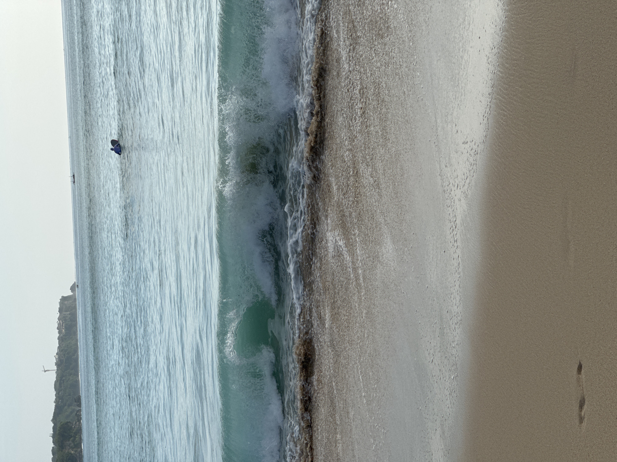 The image shows a section of beach in Kuta Selatan, Indonesia. The scene depicts the shoreline where the ocean meets the sand. There is a wave crashing onto the shore, creating white foamy water at the edge. The sand is wet near the water and drier further inland. A single person is visible far out in the ocean and near a rock and cliff. A single footprint is visible on the beach. The image is taken from an elevated angle.